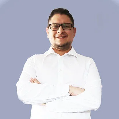 A man wearing glasses and a white shirt smiling with arms crossed against a light blue background.