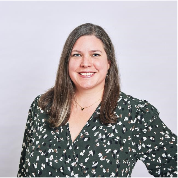 A woman with long brown hair smiling, wearing a dark patterned blouse and a small gold necklace, against a plain light background.