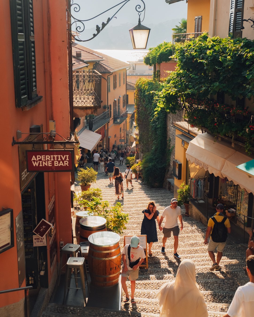People walking down a cobblestone street lined with colorful buildings, balconies with plants, and signs for a wine bar and Italian restaurant, in a scenic, sunny location.