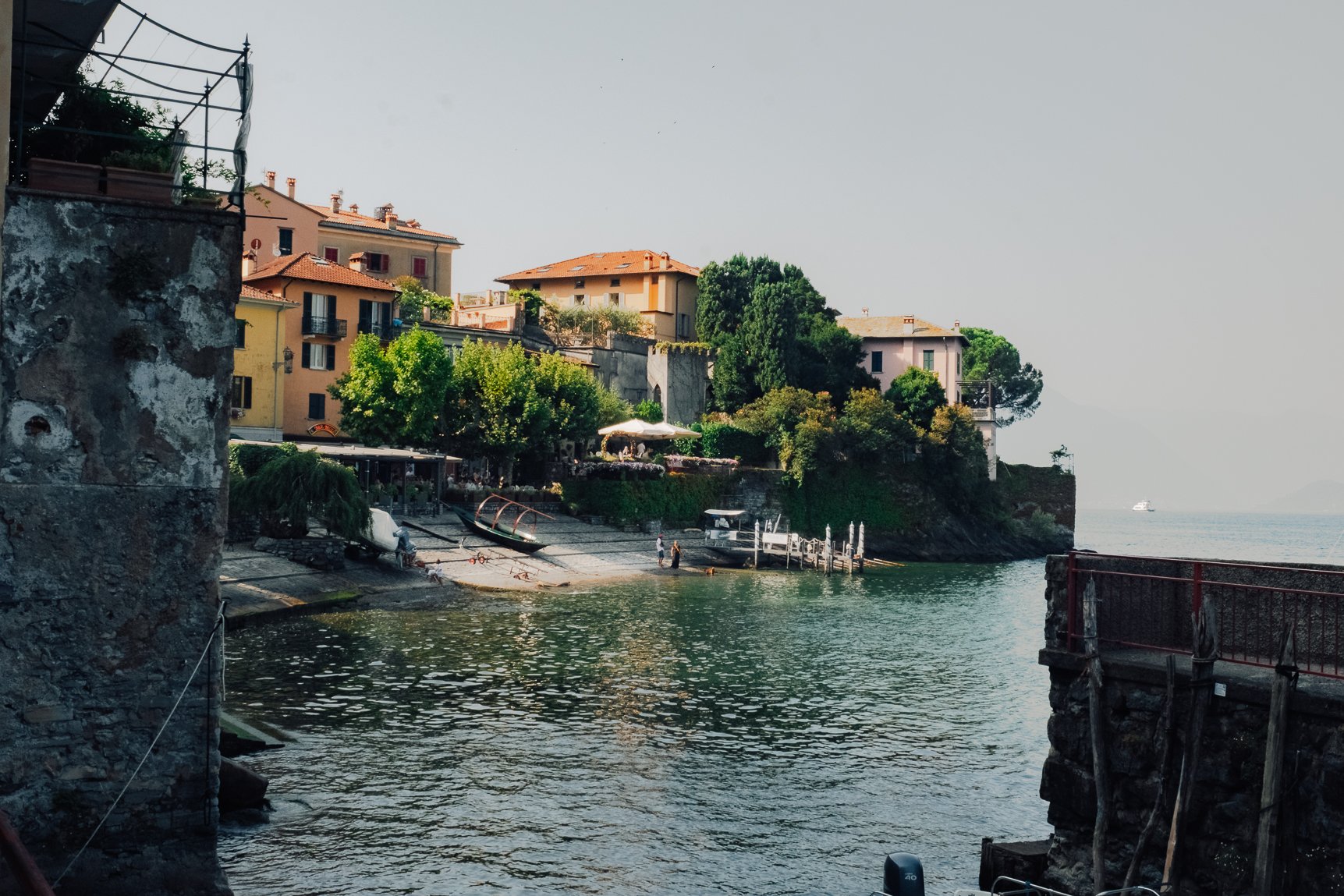 A quiet coastal harbor scene featuring historic buildings and boats framed by vibrant green trees.