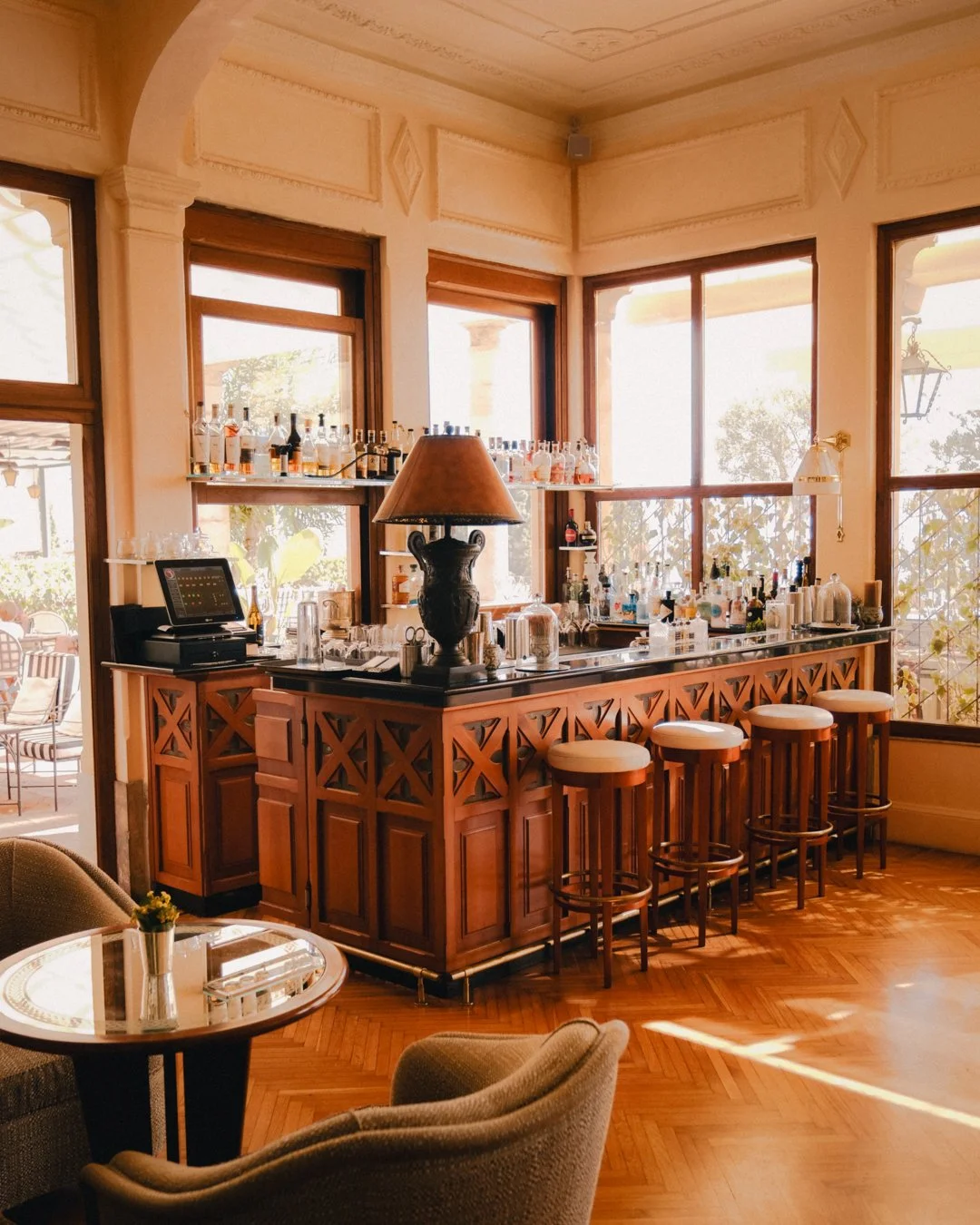 A cozy bar area with a wooden counter, bar stools, tall windows letting in natural light, and shelves with various bottles of liquor, in a warmly lit room with hardwood floors and elegant decor.