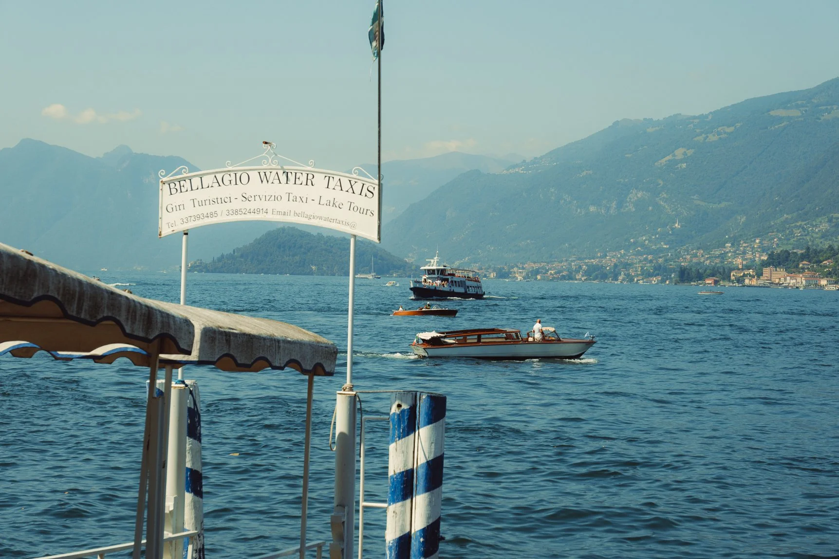 Classic view of a Lake Como ferry dock with blue and white striped poles and mountains in the distance.