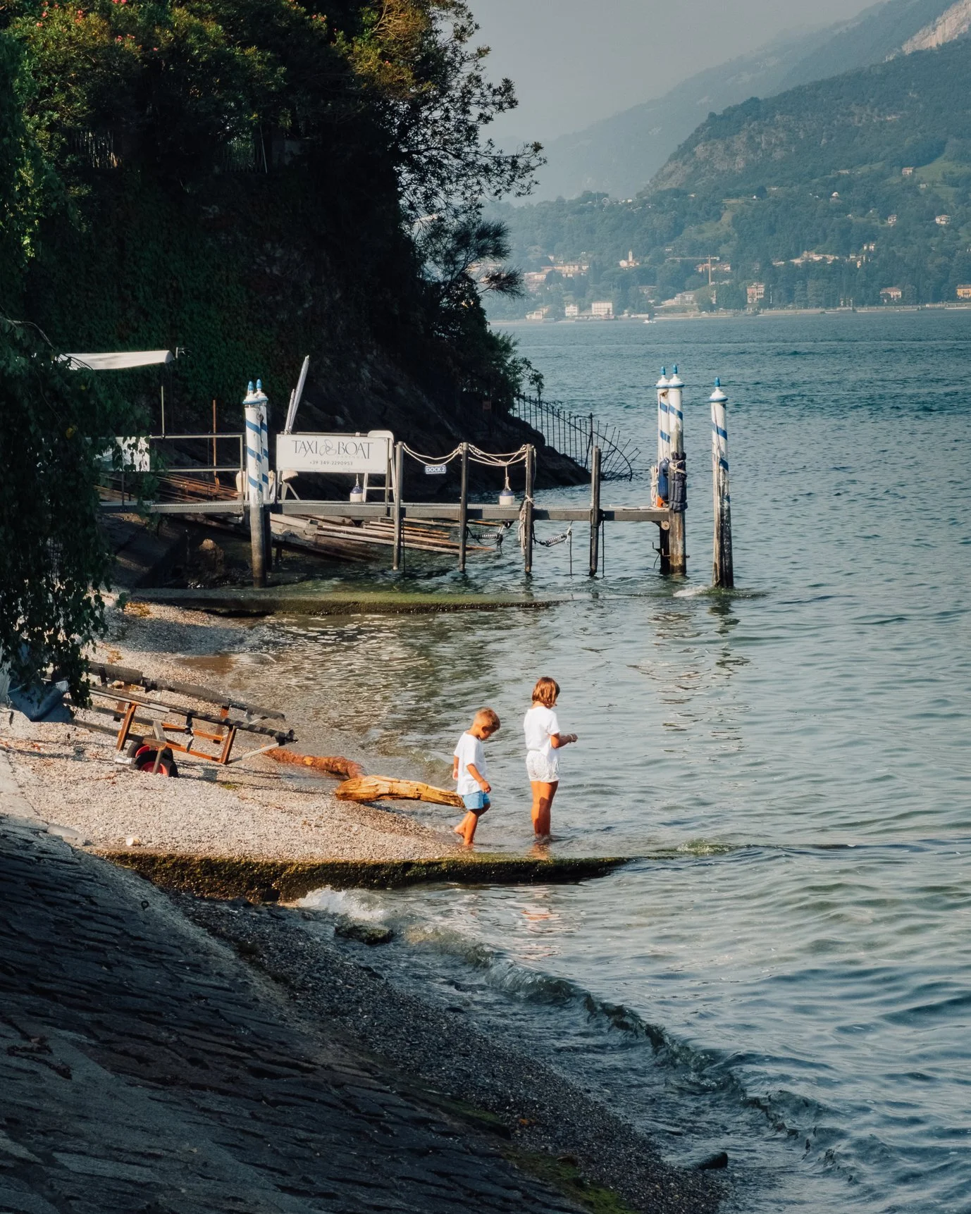 Children and swimmers playing on a wooden dock by the lake edge, bathed in warm afternoon light.