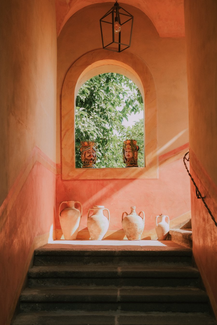 Indoor stairwell with terracotta-colored walls leading up to a small landing with four ancient-style terracotta vases and two masks on the window ledge, surrounded by greenery outside.