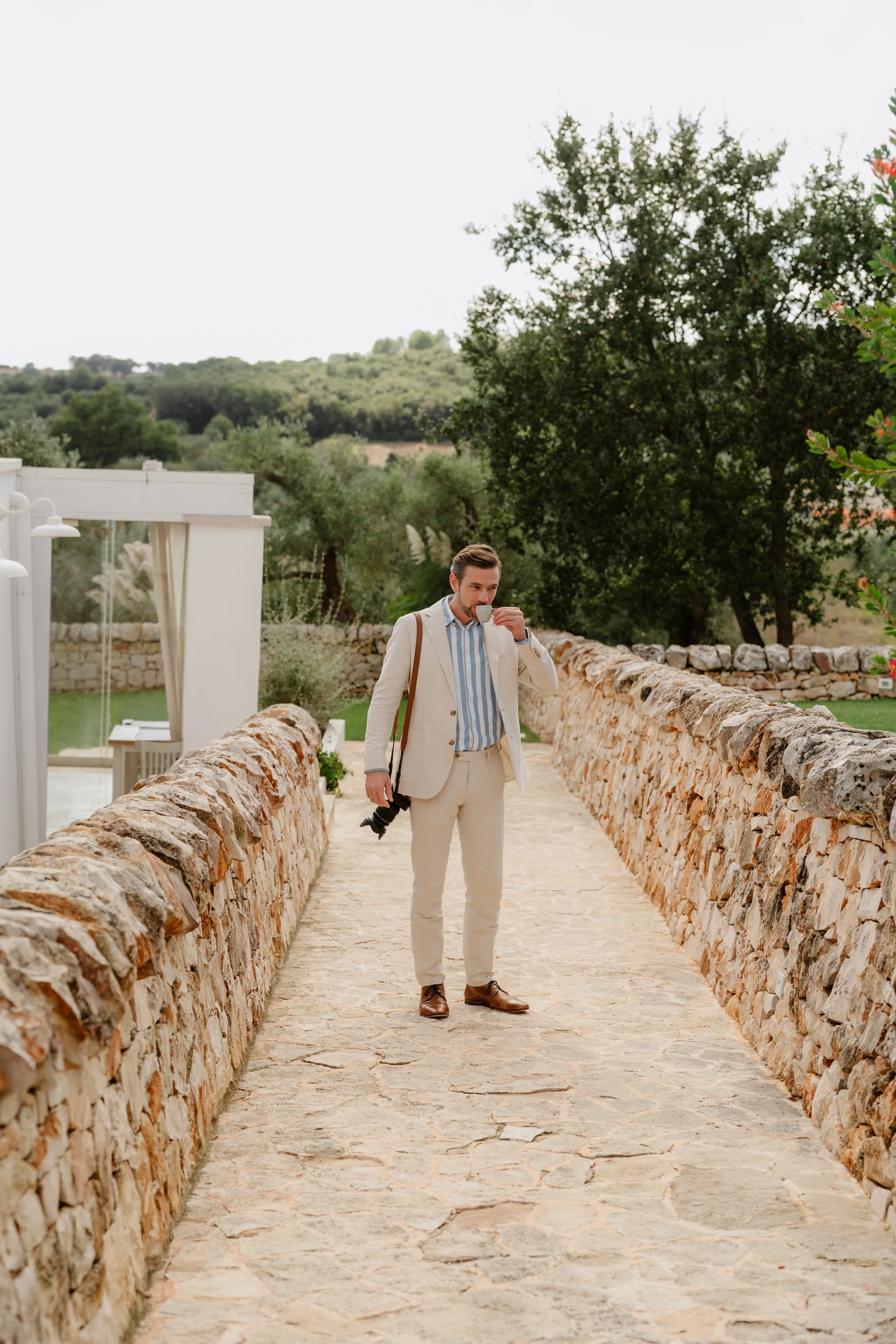 A man wearing a cream-colored suit, striped shirt, and brown shoes walking on a stone pathway while drinking from a cup of coffee, carrying a camera and brown shoulder bag, with stone walls and green trees in the background.