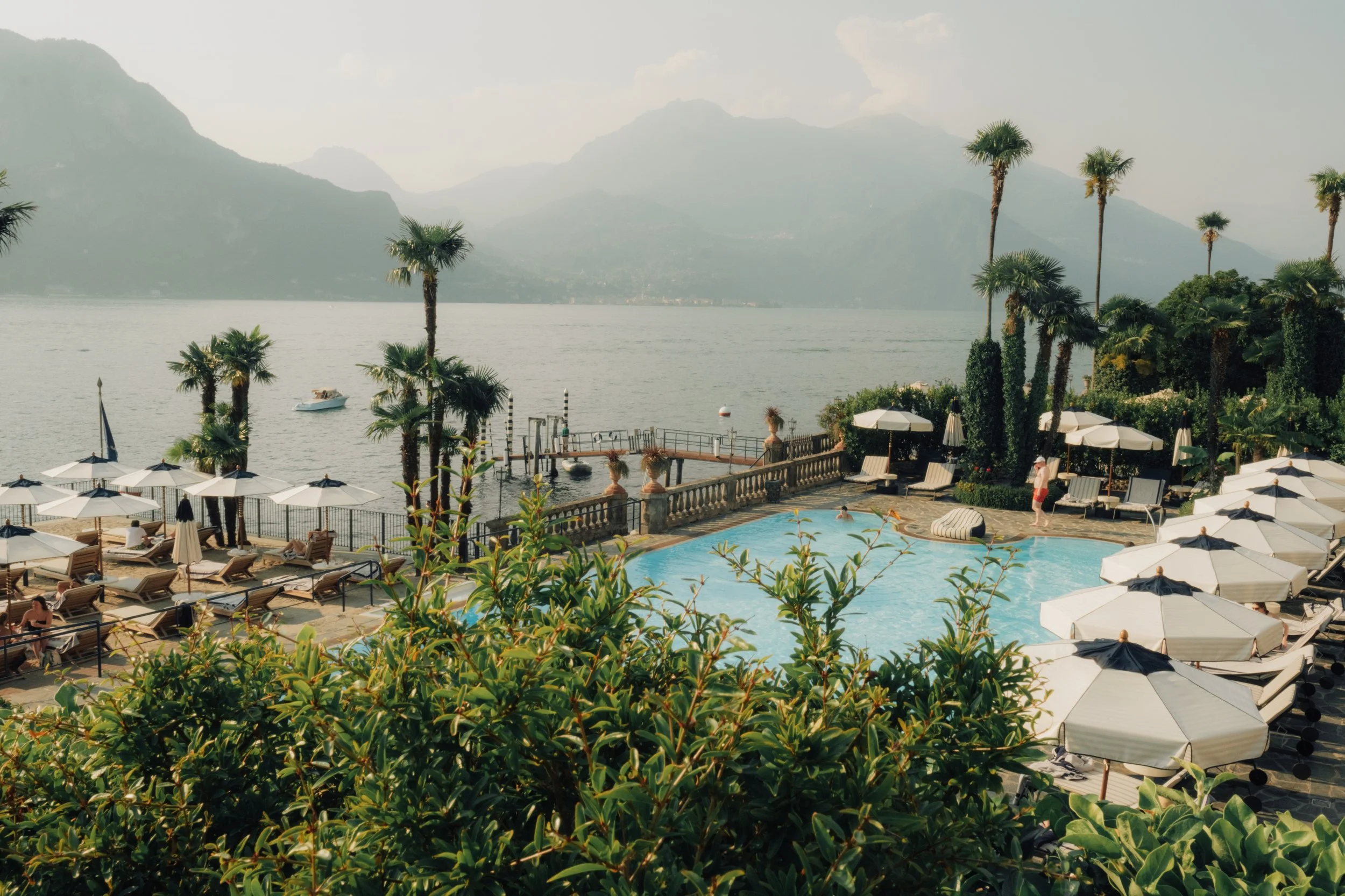A serene hotel pool area framed by cypress trees and mountains, capturing a quiet morning atmosphere.