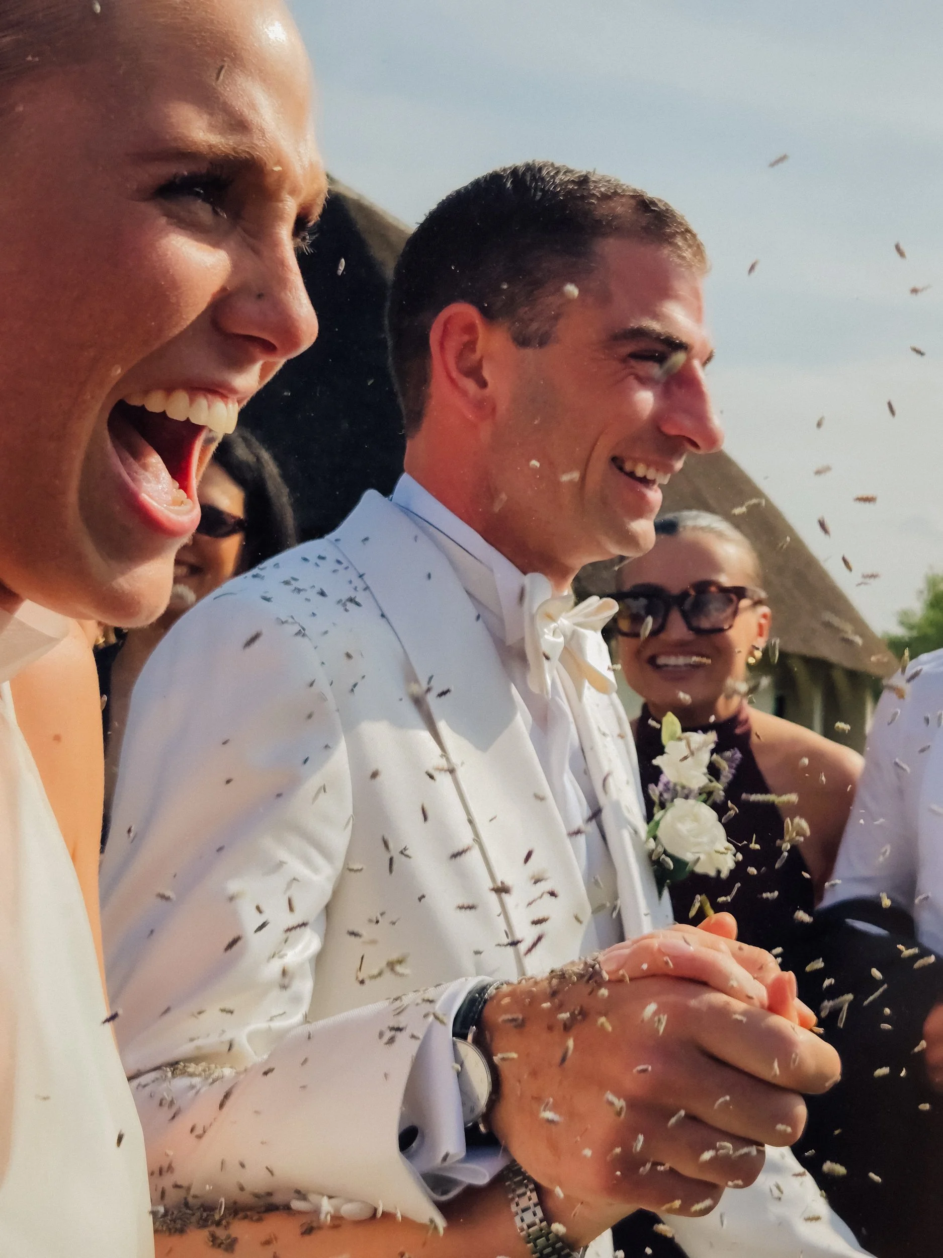 A group of people celebrating outdoors, including a man in a white tuxedo with a bow tie, a woman in a white dress, and others smiling as they throw confetti.