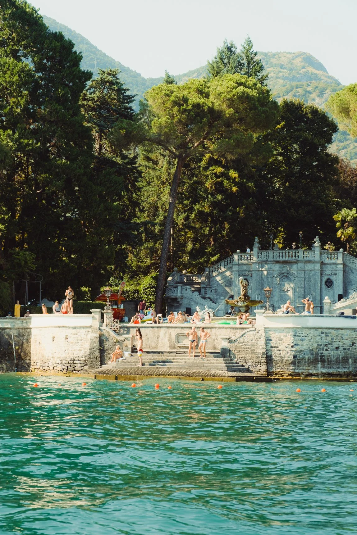 People relaxing on white lounge chairs by a swimming pool surrounded by tall, lush palm trees and greenery.