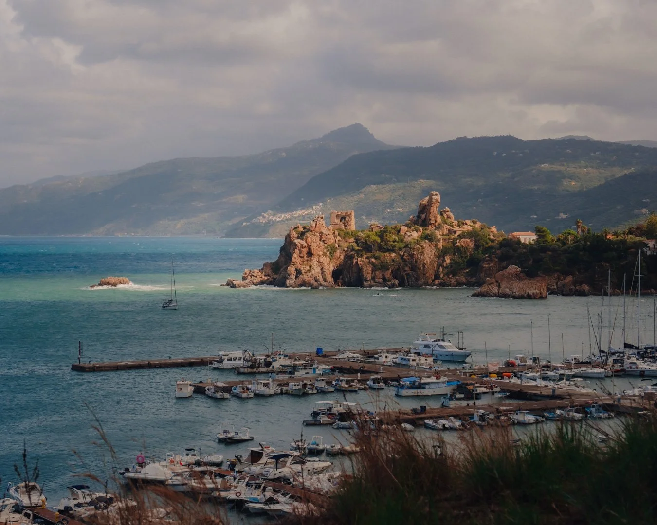 A harbor with numerous boats and yachts docked along piers, a rocky cliff with ruins and greenery, a calm sea, and a mountain range in the background under a partly cloudy sky.