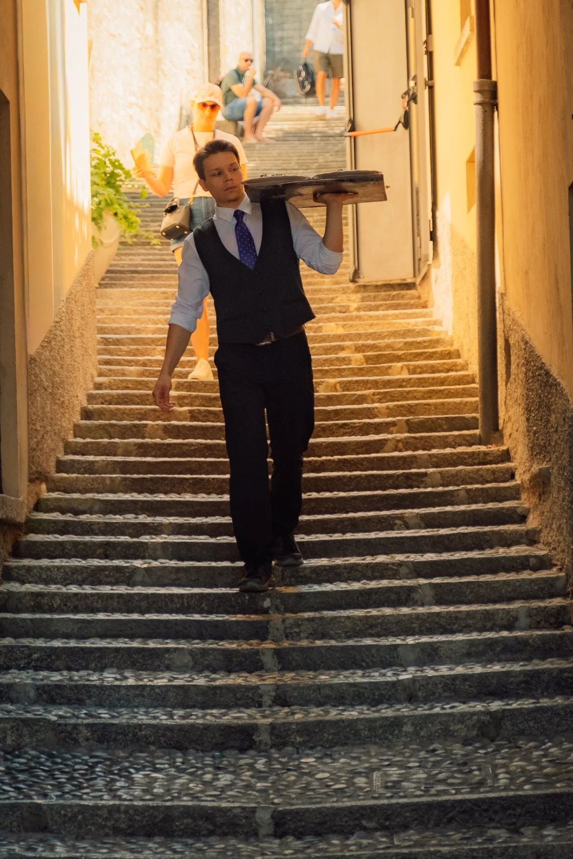 A waiter in a vest carrying a silver tray down sun-drenched stone steps in an Italian village.