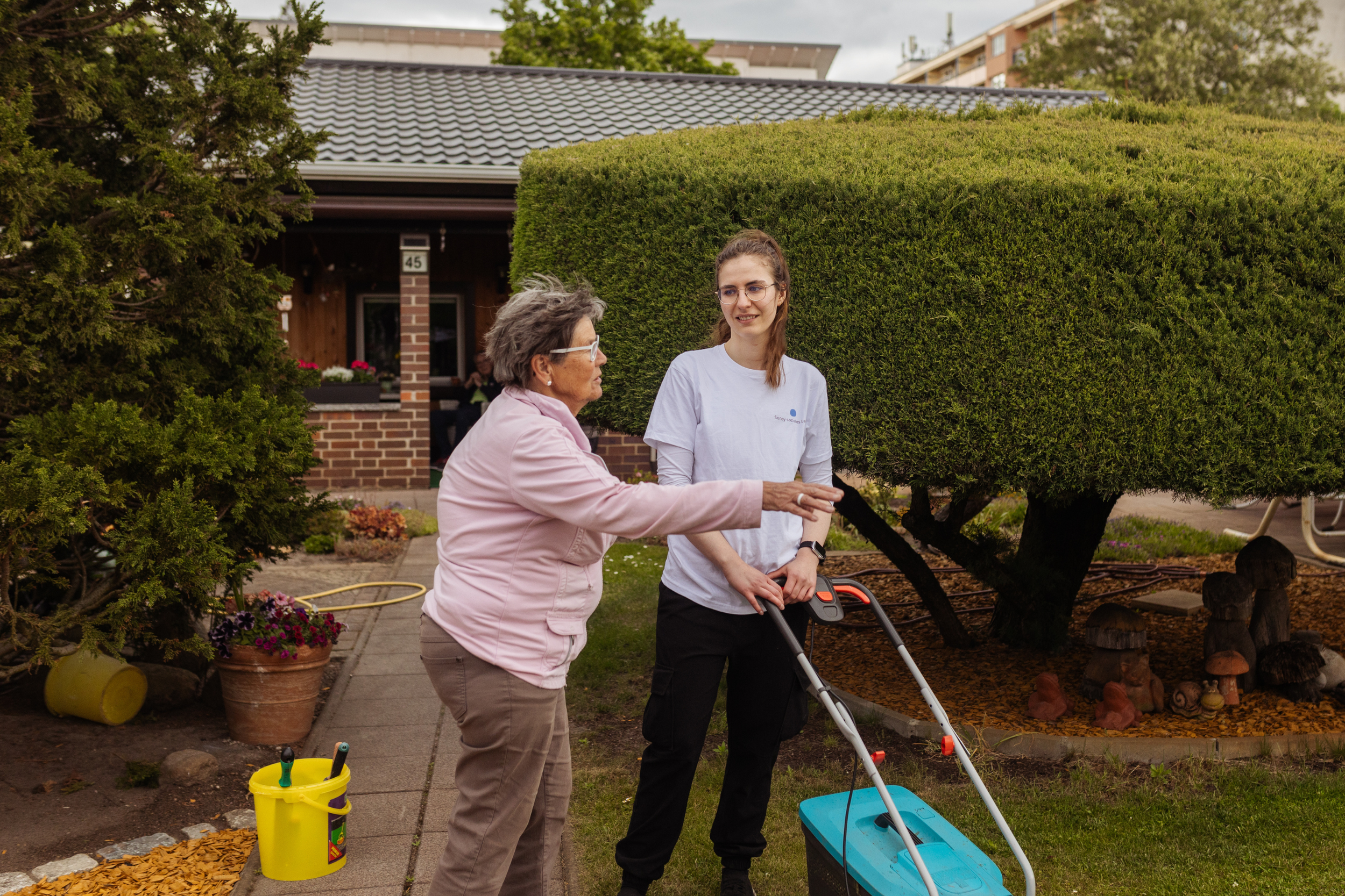 Eine ältere Frau und eine jüngere Frau  Sonay soziales Leben e.V. im Projekt Teilhabe-Hilfe-Card Jugendliche und Senioren sprechen im Garten. Die jüngere Frau hält eine Rasenmäher.]