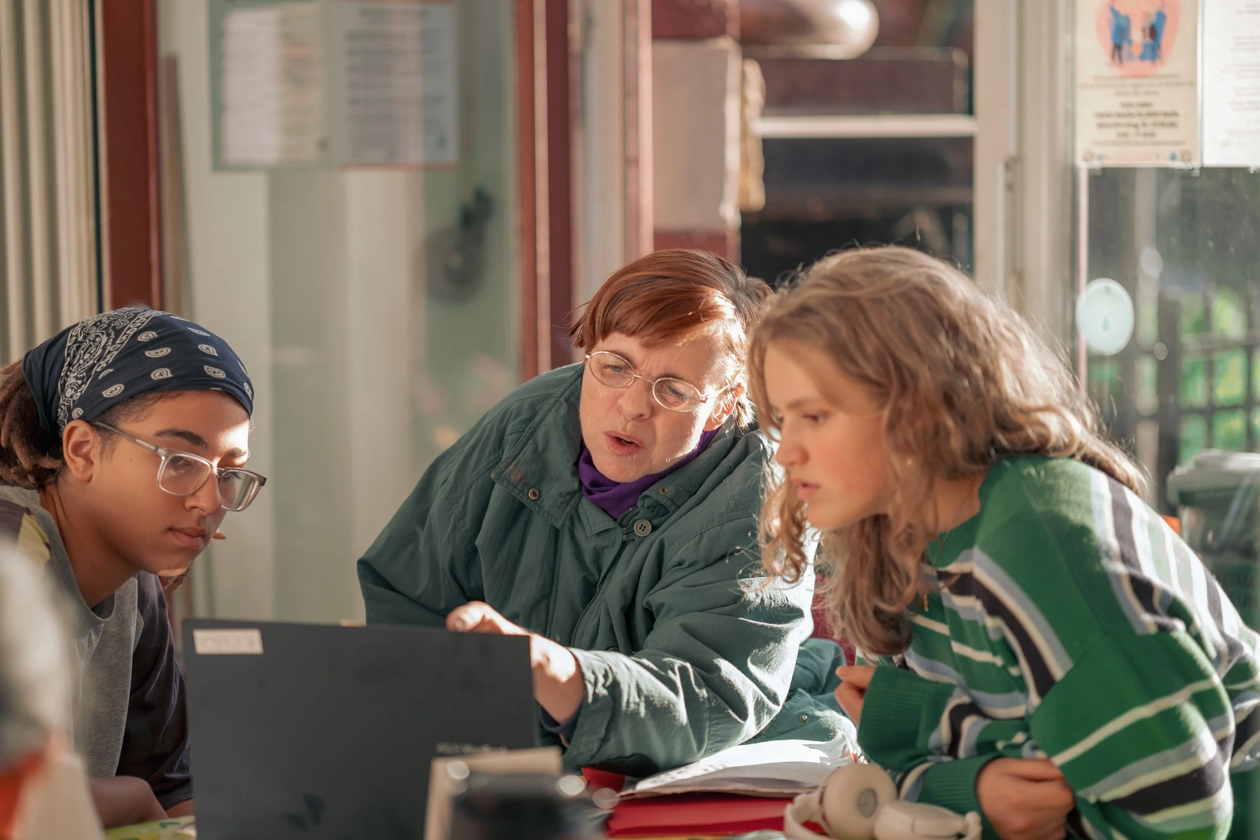 Drei Menschen  Sonay soziales Leben e.V. im Projekt Teilhabe-Hilfe-Card Jugendliche und Senioren, ein Mädchen, eine Frau und eine ältere Frau, schauen auf einen Laptop in einem Raum mit großen Fenstern und verschiedenen Papieren an der Wand.