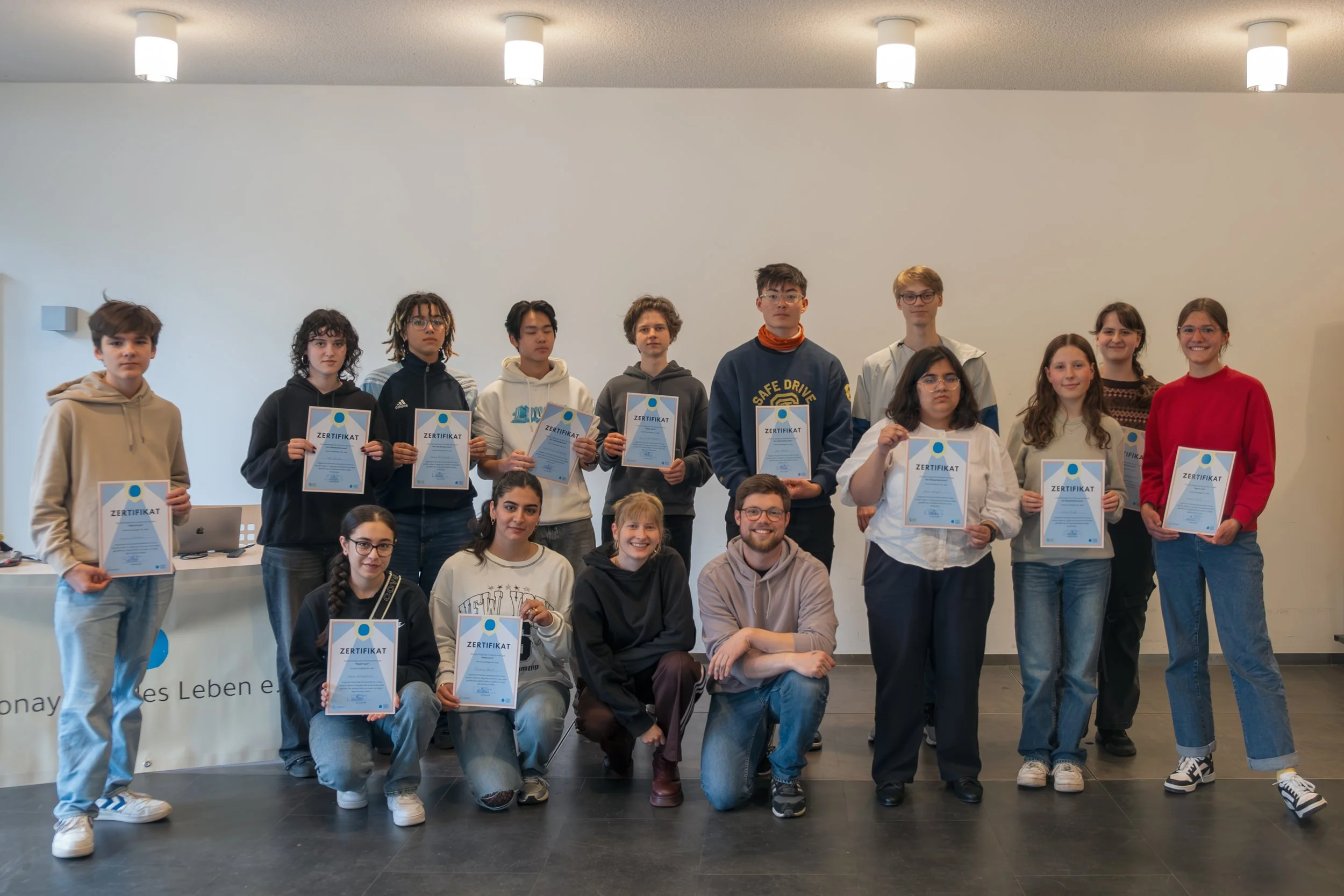 Gruppe Jugendlicher  Sonay soziales Leben e.V. im Projekt Teilhabe-Hilfe-Card Jugendliche und Senioren, die Zertifikate in einem Raum halten, vermutlich bei einer Preisverleihung oder Zertifikatsübergabe, alle lächeln und schauen in die Kamera.