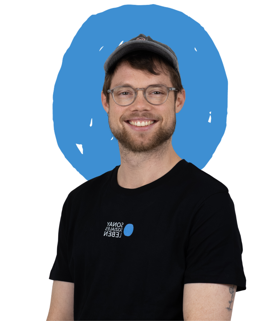 Ein junger Mann  Sonay soziales Leben e.V. im Projekt Teilhabe-Hilfe-Card Jugendliche und Senioren mit Brille, Bart und einem schwarzen T-Shirt, das den Satz „Sony Dasgen Leben“ mit einem blauen Punkt zeigt, lächelt vor einem blauen Hintergrund.