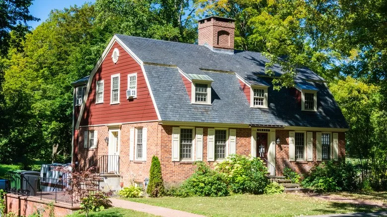 Red brick house with a dark gray shingled roof, multiple windows, and a chimney, surrounded by green trees and a well-maintained lawn.