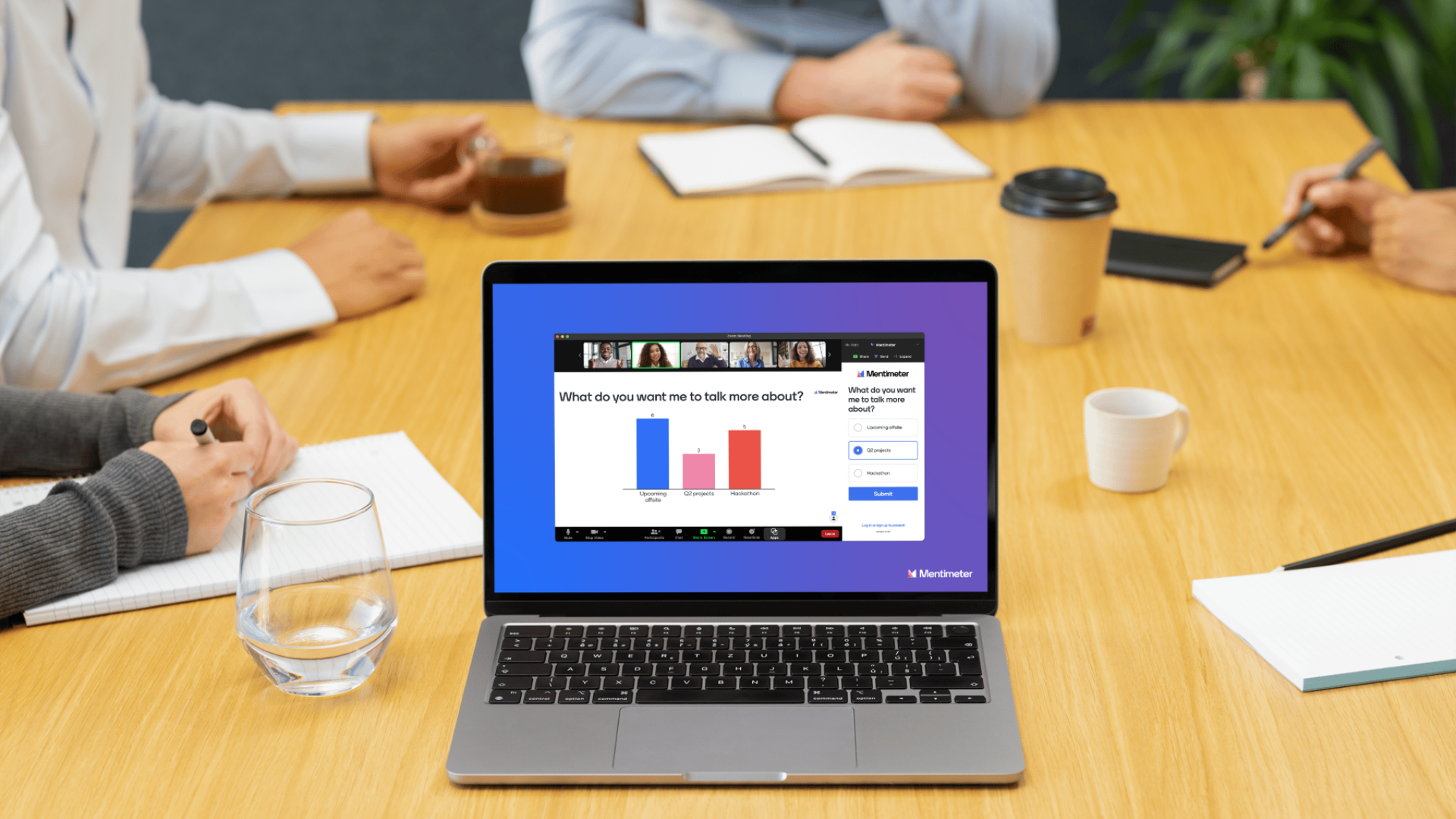 A laptop on a conference table displaying a virtual meeting poll about topics to discuss, surrounded by notebooks, coffee cups, a glass of water, and people taking notes.