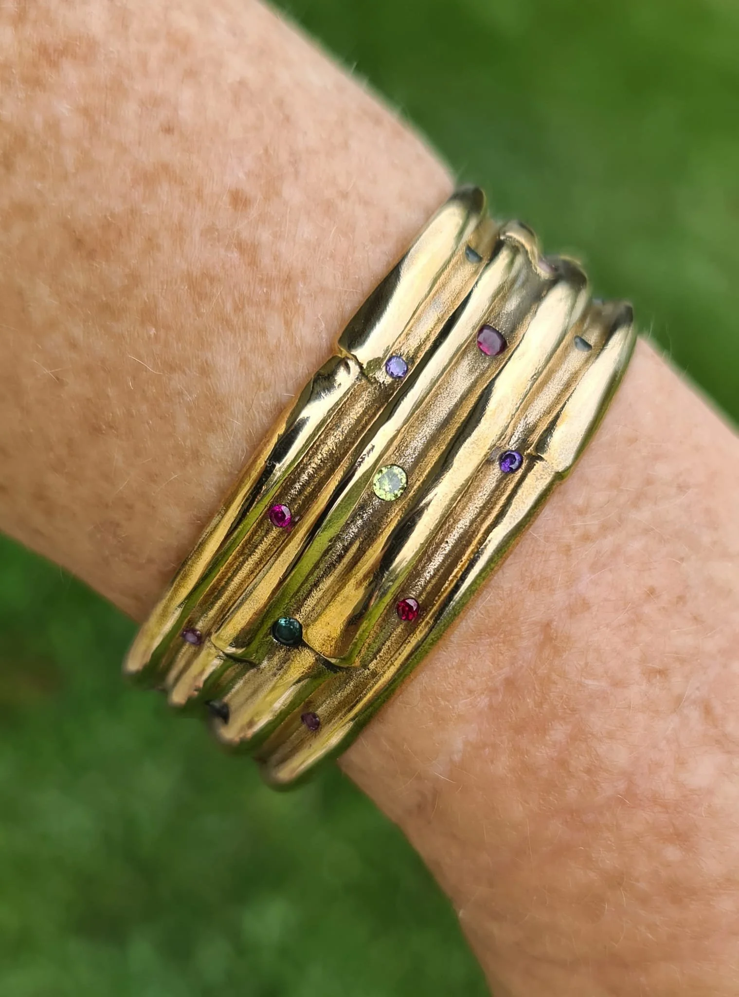 Close-up of multiple gold bangles adorned with small colorful gemstones on a person's wrist, with a blurred green background.