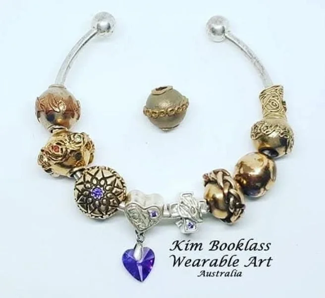 A silver bracelet with gold and silver charms and a purple heart pendant, accompanied by a matching charm. The jewelry is displayed on a white background with a label that reads "Kim Booklass Wearable Art Australia."