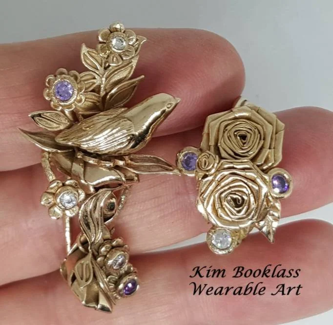 Gold jewelry accessory with a bird, roses, leaves, and small purple and clear gemstones, worn on a finger.
