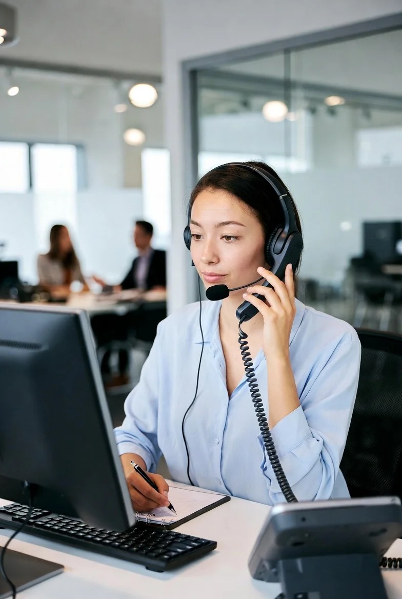 A young woman in a blue shirt wearing a headset with microphone, working at a computer in an office with other people in the background.