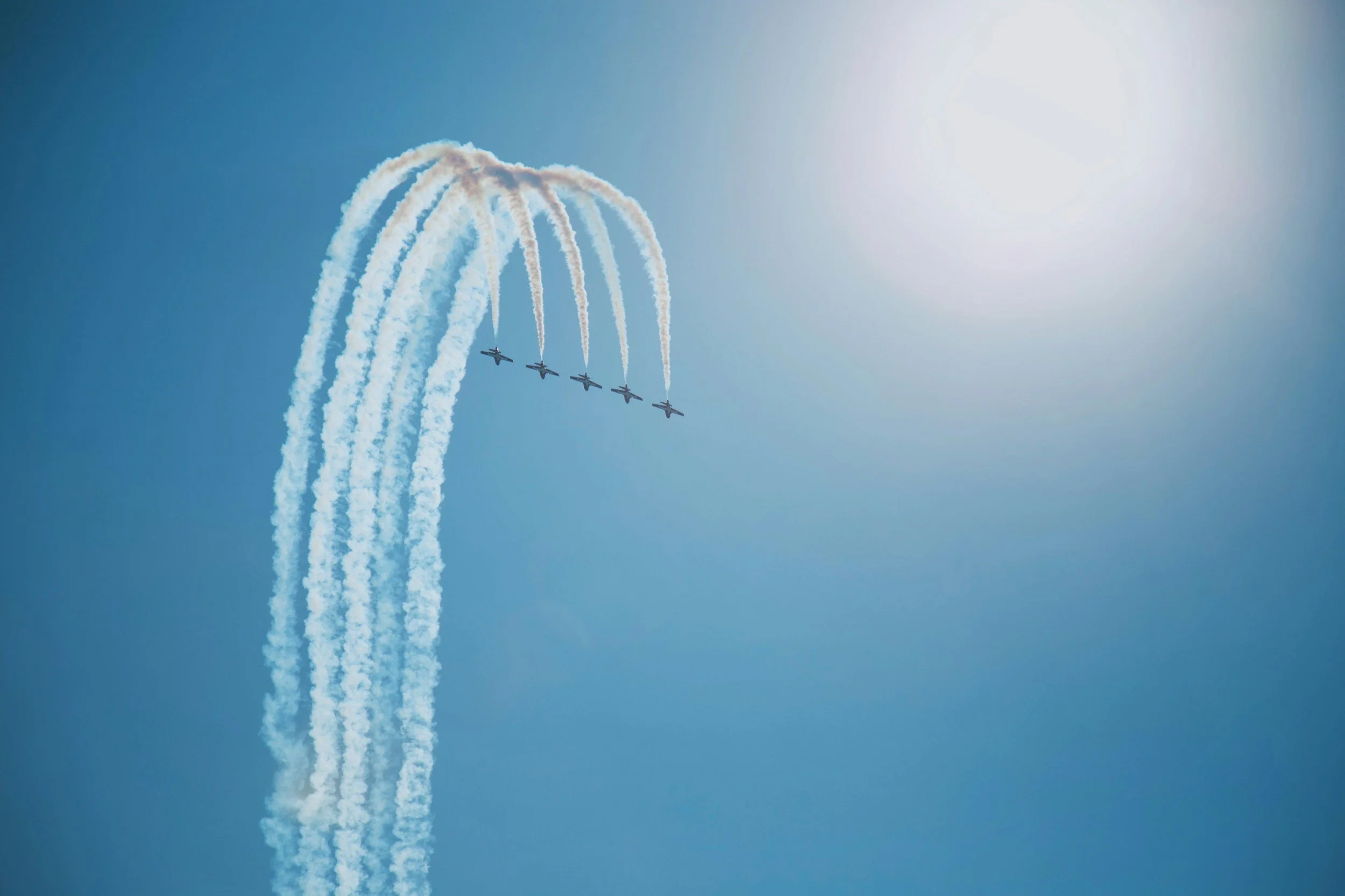 Five airplanes flying in a formation, leaving white smoke trails against a bright blue sky with the sun shining.