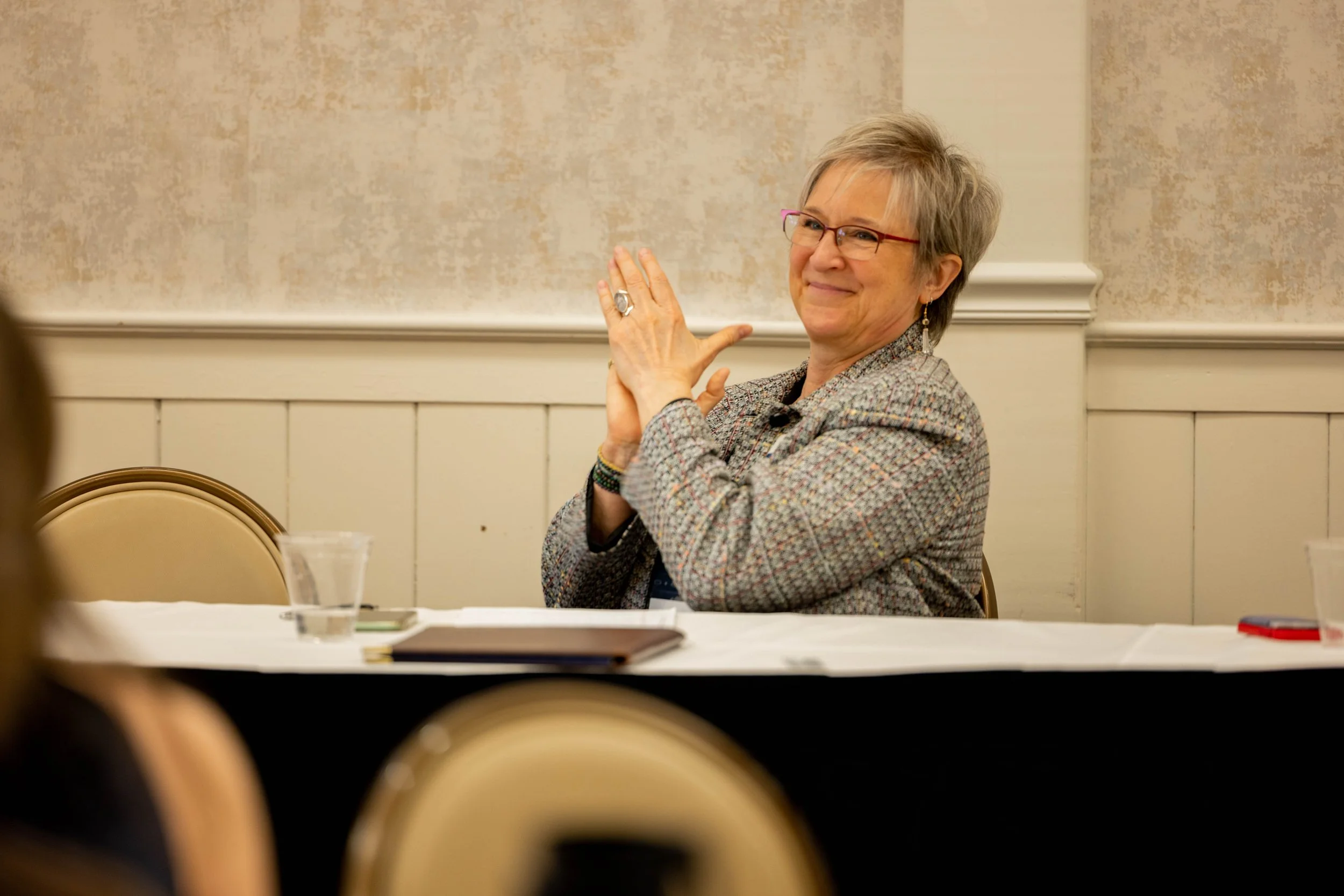 A smiling woman with short gray hair, glasses, and earrings sitting at a conference table, gesturing with her hands during a meeting or presentation.