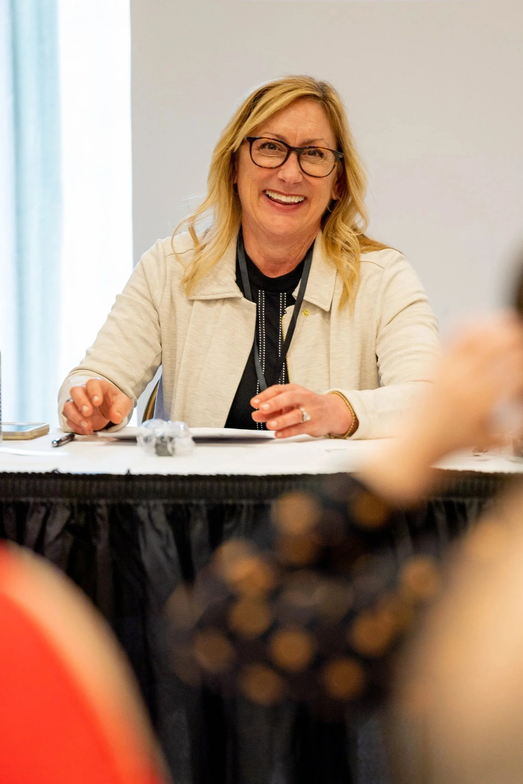 A smiling woman with blonde hair and glasses sitting at a table during a meeting or conference.
