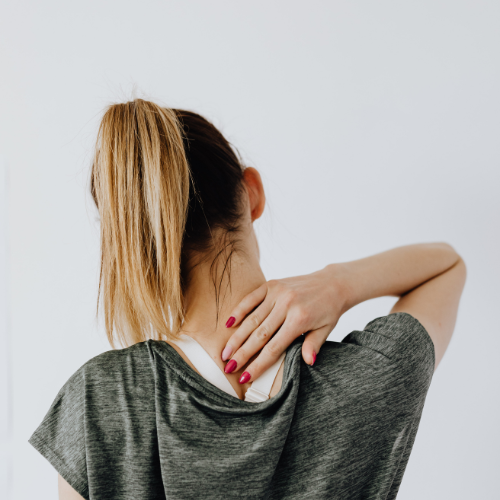 Woman with blonde hair in a ponytail scratching her neck with her right hand, wearing a gray shirt against a white background.