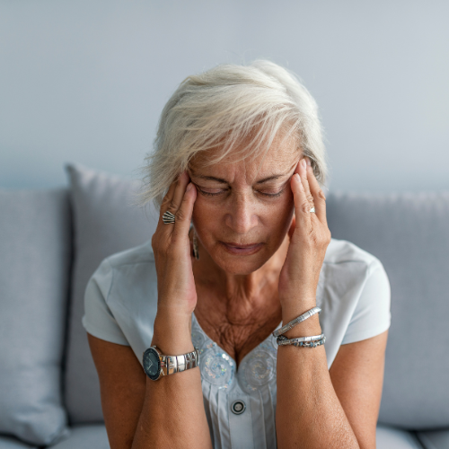 An elderly woman with short white hair sitting on a couch, holding her head with both hands, appearing distressed or in pain.
