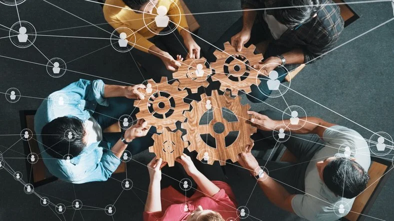 Top-down view of five people working together around a wooden table with large gear-shaped cutouts, overlaid with digital network icons and lines.