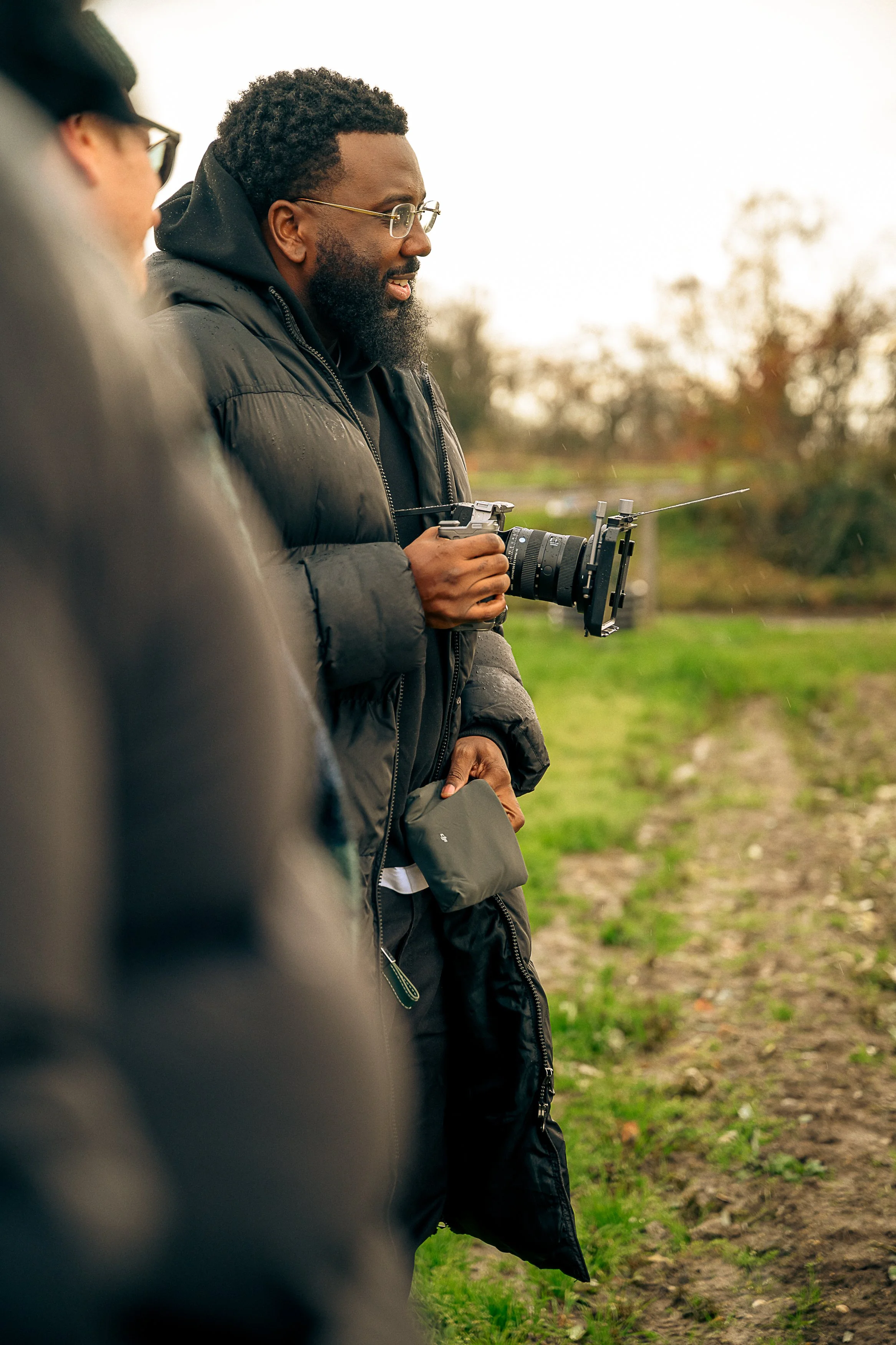 A man with glasses, a black puffer jacket, and a beard holding a professional camera with a stabilizer in a rural outdoor setting, with blurred trees and fields in the background.