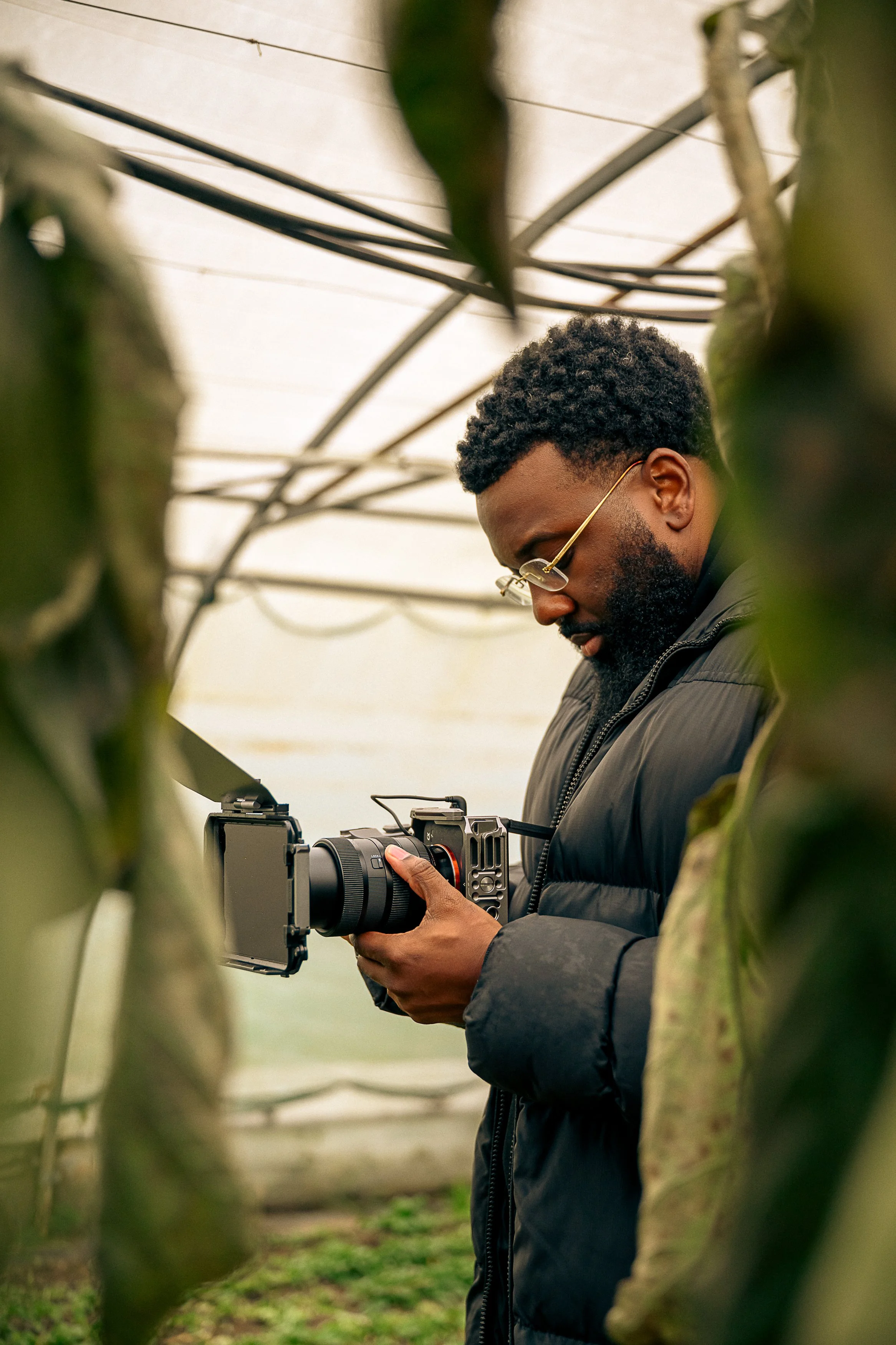 A man with curly hair and a beard wearing glasses and a black jacket, holding a camera inside a greenhouse or plant tunnel with green plants and a cloudy sky overhead.
