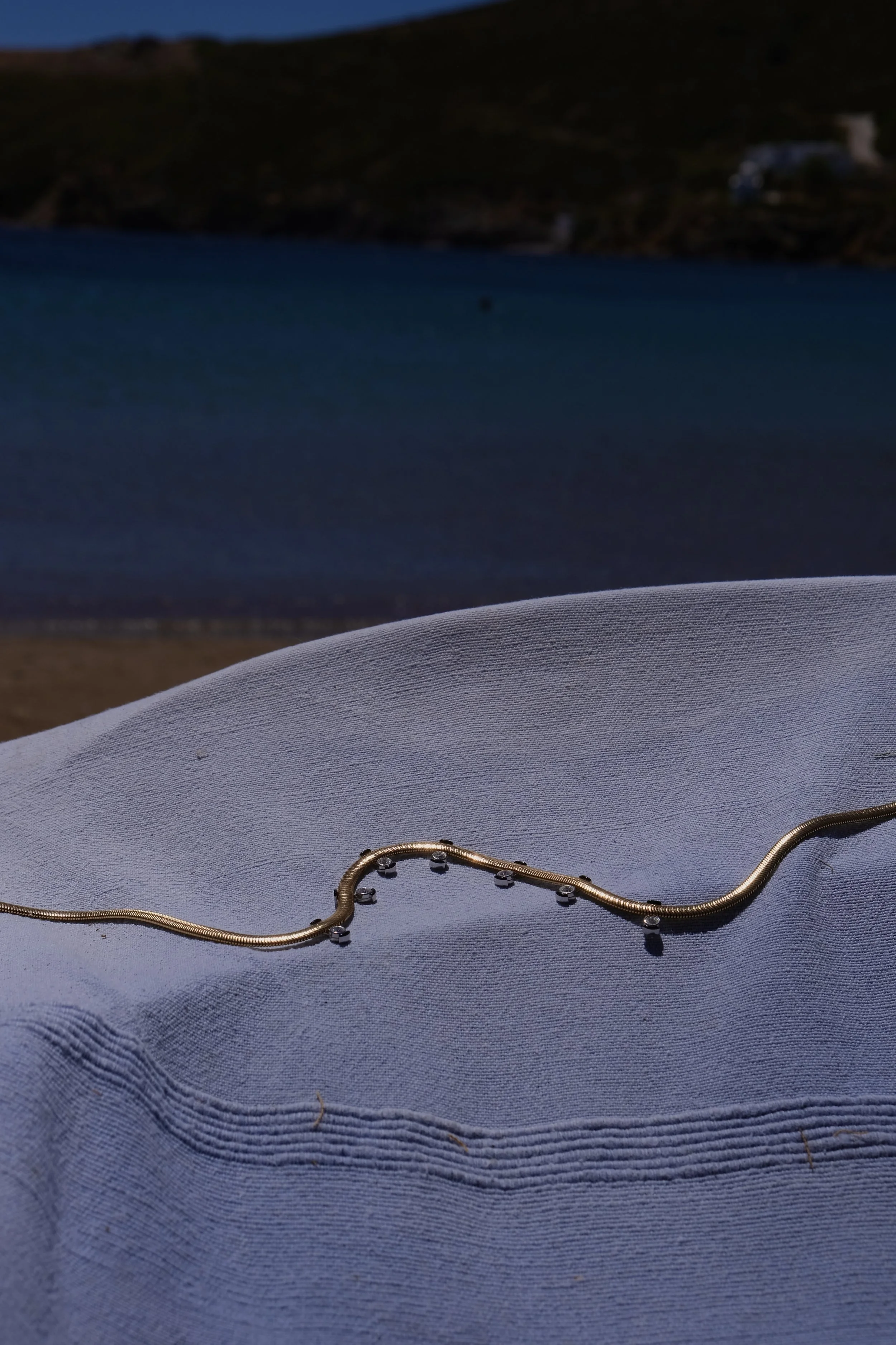 A silver necklace with small beads resting on a folded blue fabric with a beach and ocean in the background.