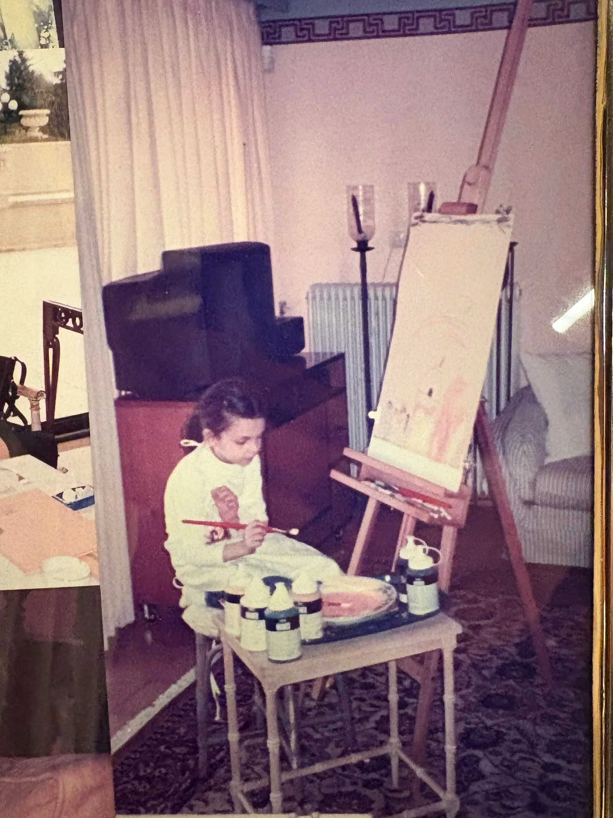 A young girl sitting on a chair painting on an easel in a room, surrounded by art supplies and furniture.