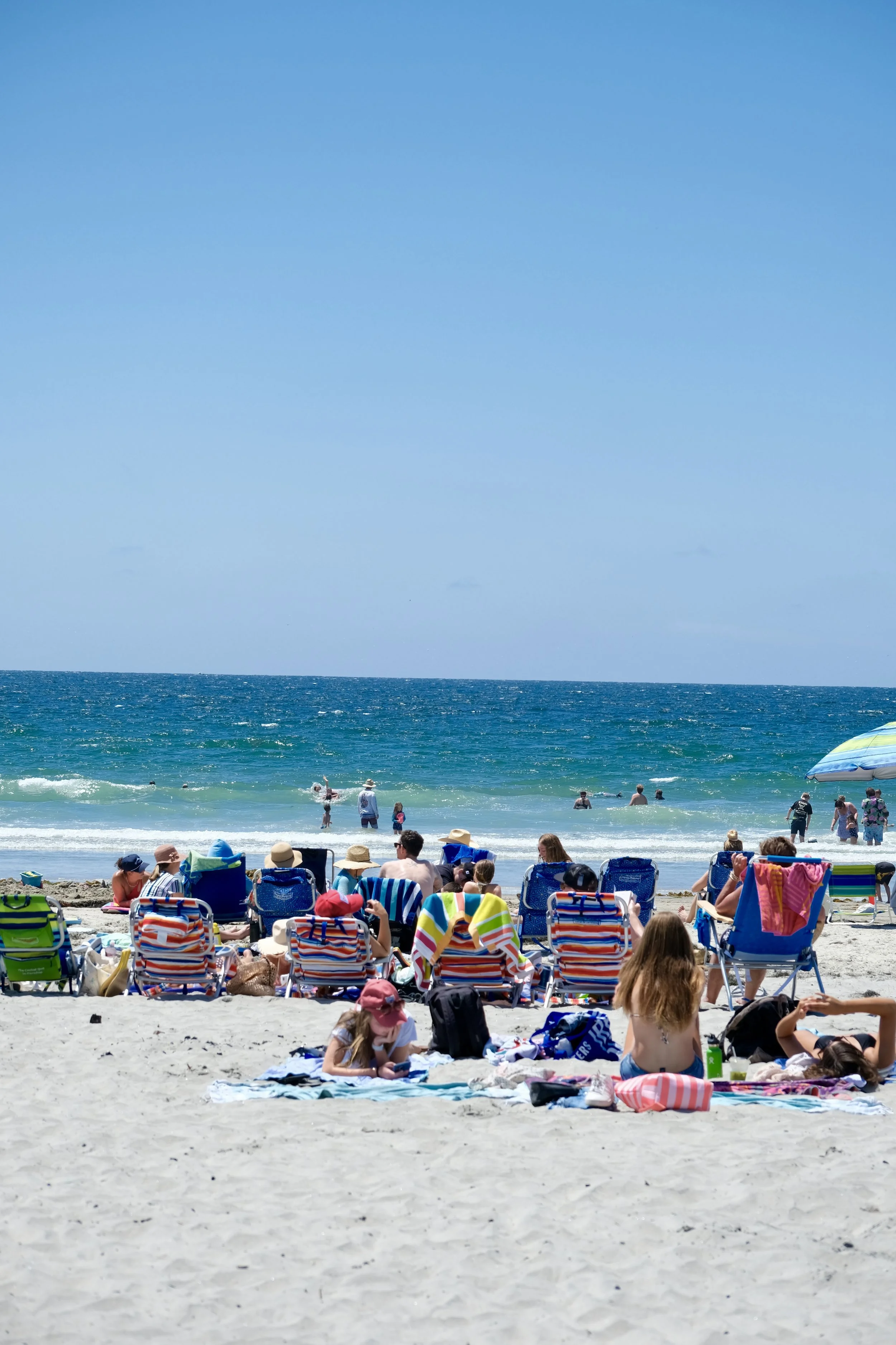 People relaxing on a sandy beach with colorful chairs and umbrellas, swimming and playing in the ocean under a clear blue sky.