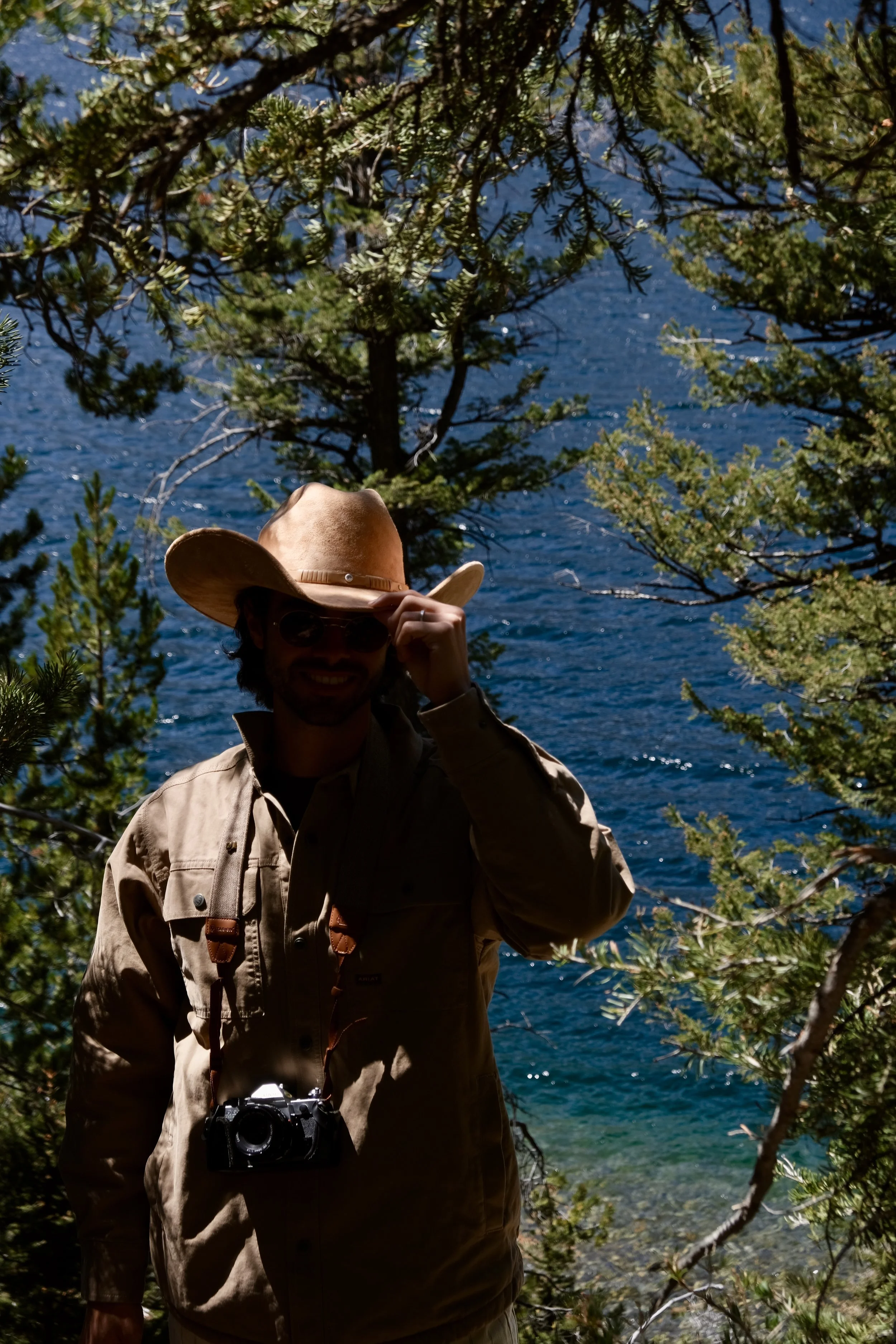 A man wearing a cowboy hat, sunglasses, and a beige jacket with a camera hanging around his neck, standing outdoors among trees near a body of water.