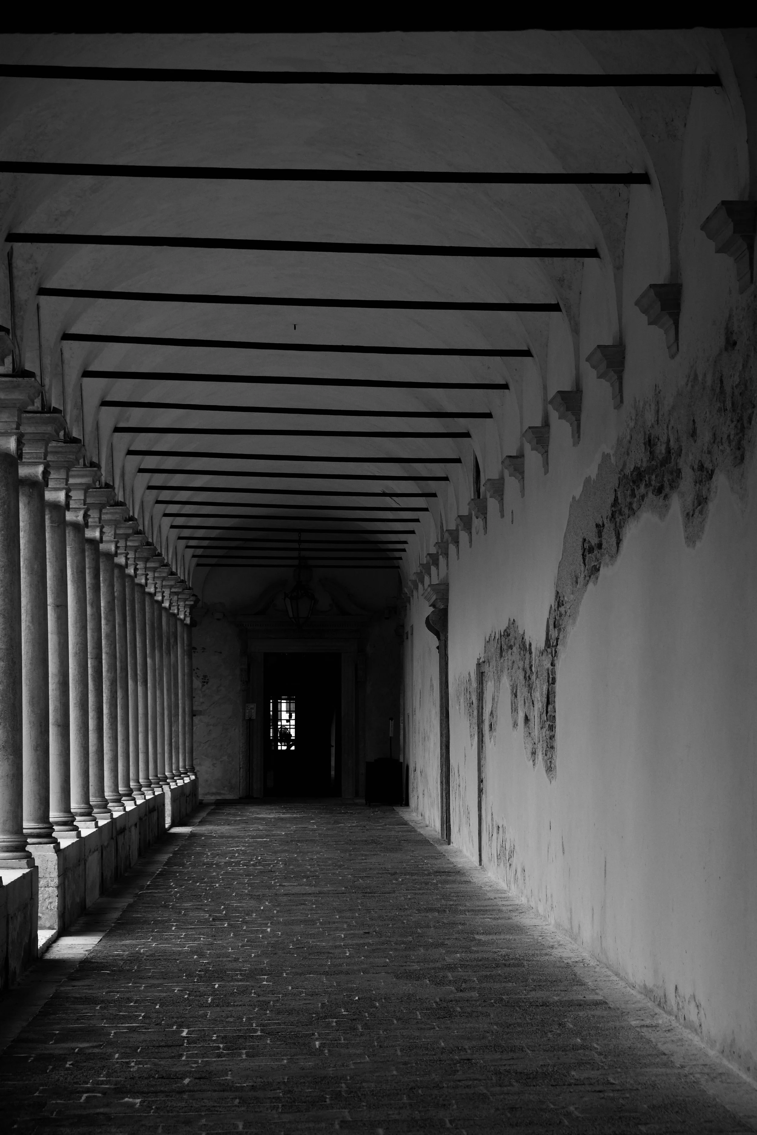 Black and white photograph of a long, arched corridor with columns on the left and a peeling wall on the right, leading to a doorway in the distance.
