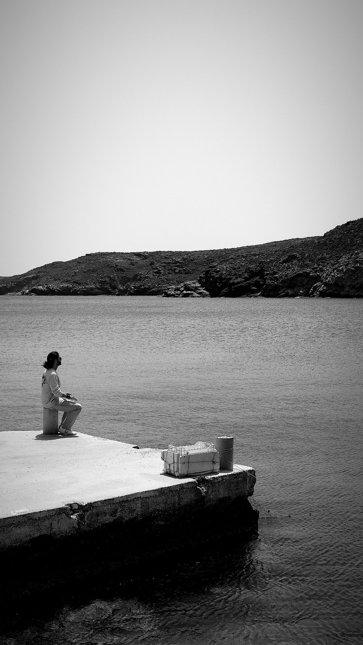 A person sitting on a small stool on a pier by the water, with a mountainous landscape in the background. The image is in black and white.