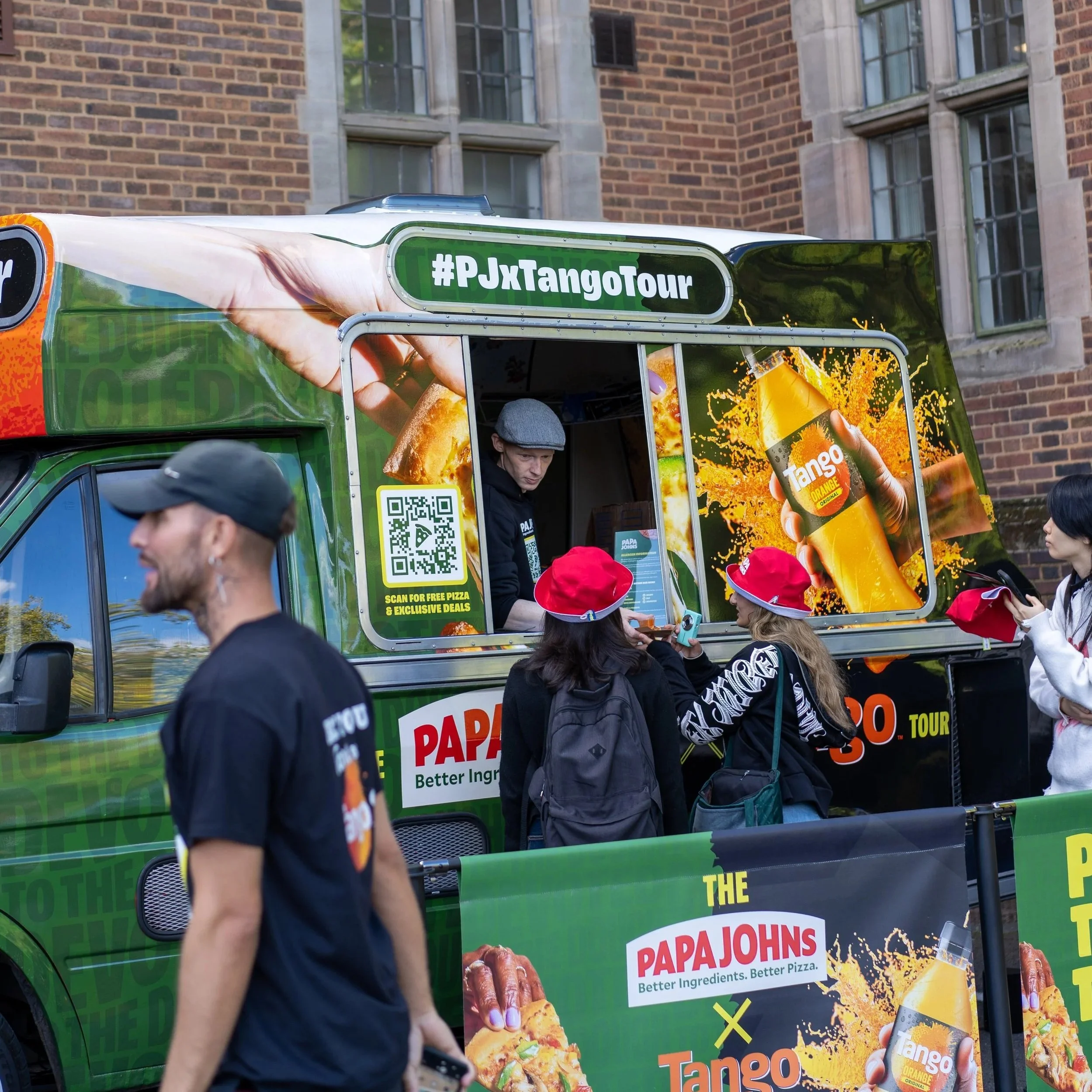 A Panera Bread food truck during a promotional event with a large sandwich and beverage advertisement. Several people with red hats are interacting with the truck, and a man in a gray cap is working inside the truck.