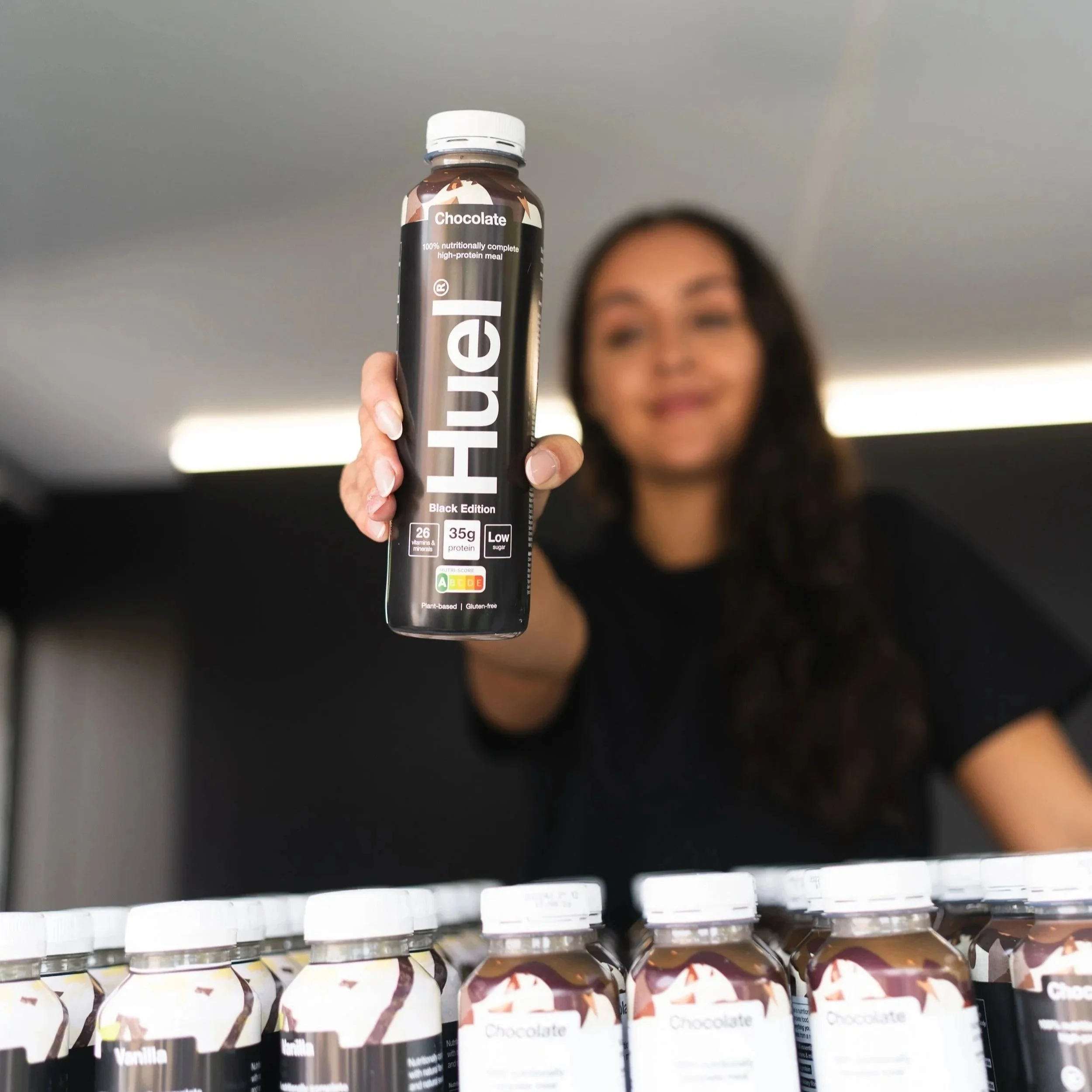 A woman holding a bottle of Huel Black Edition Chocolate flavor in front of a row of similar bottles.