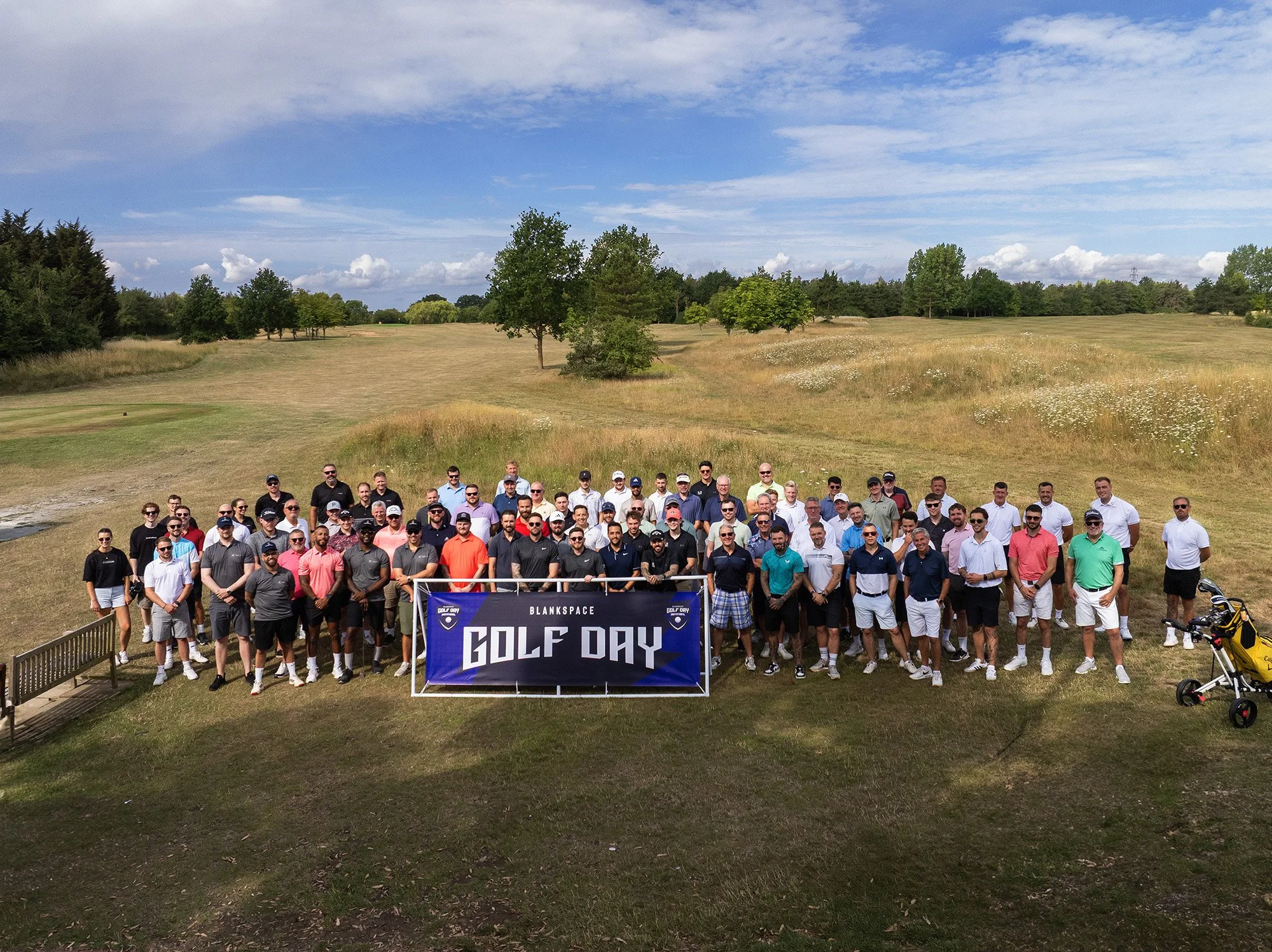 Group of golf players posing on a golf course holding a banner that reads 'GOLF DAY' under a partly cloudy sky.
