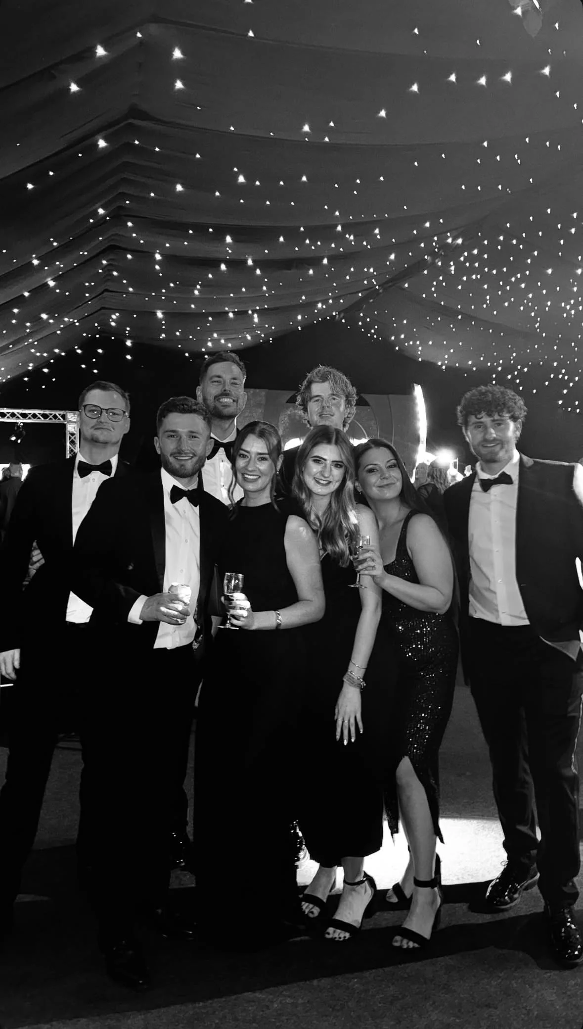 Group of people at a formal event, dressed in tuxedos and evening gowns, under a ceiling decorated with string lights, smiling and posing for the camera.