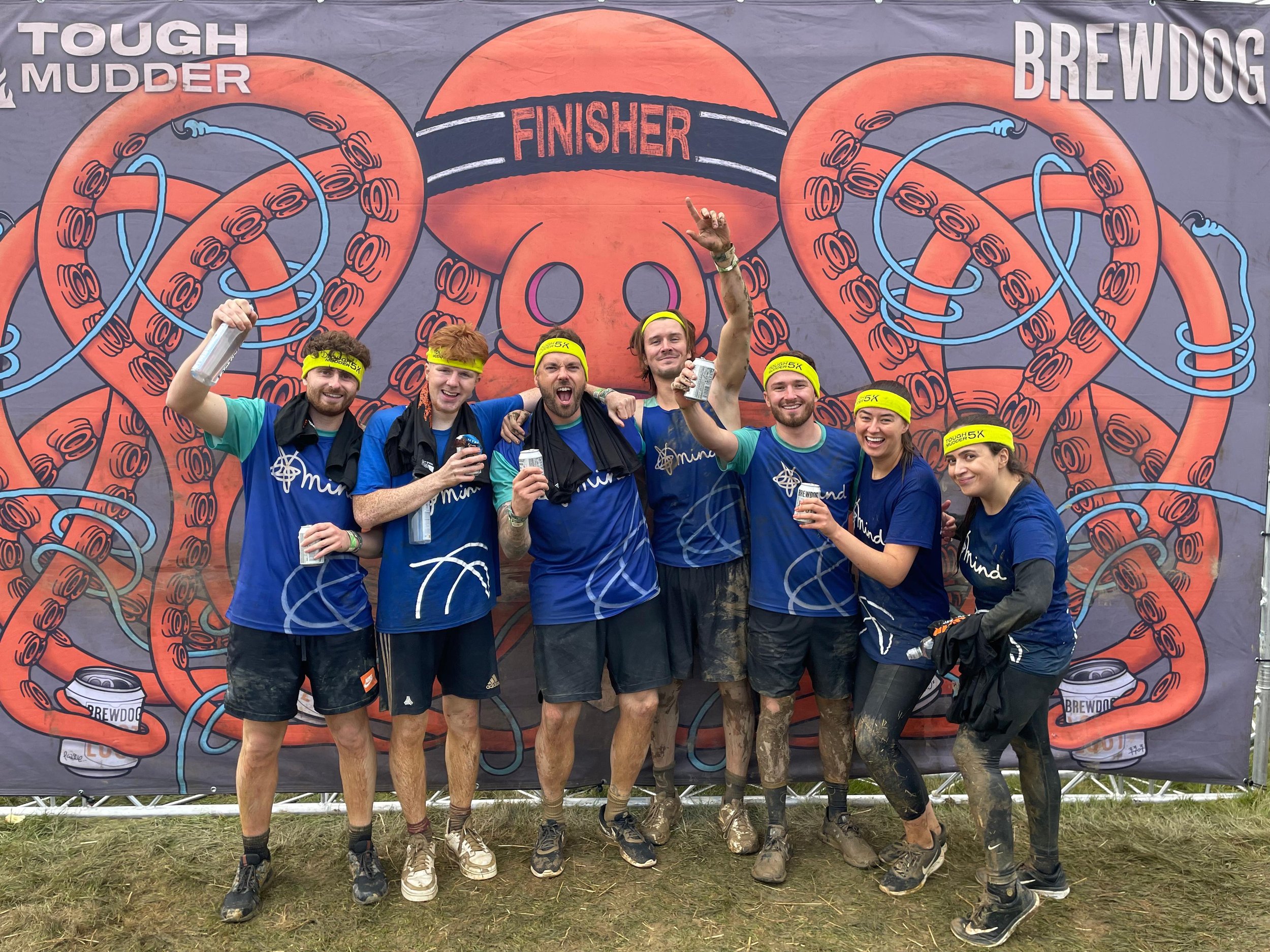 A group of seven people in blue shirts and yellow headbands celebrating their finish at the Tough Mudder and Brewdog event. They are posing in front of a large backdrop with cartoon-style tentacles and the words 'Finisher' and 'Tough Mudder' and 'Brewdog.' They are holding drinks, smiling, and some have mud on their legs.