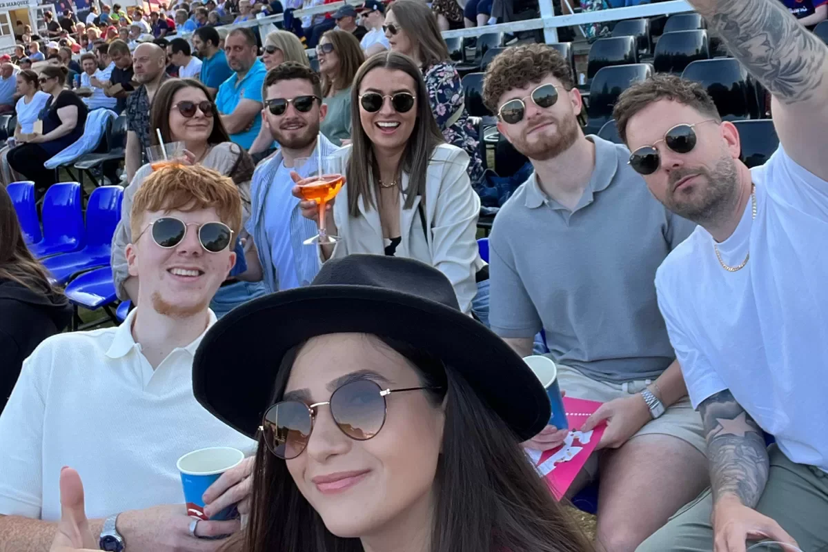 A group of friends sitting in a stadium, smiling at the camera while taking a selfie. They are wearing sunglasses, and some are holding drinks. The crowd and empty seats can be seen in the background.