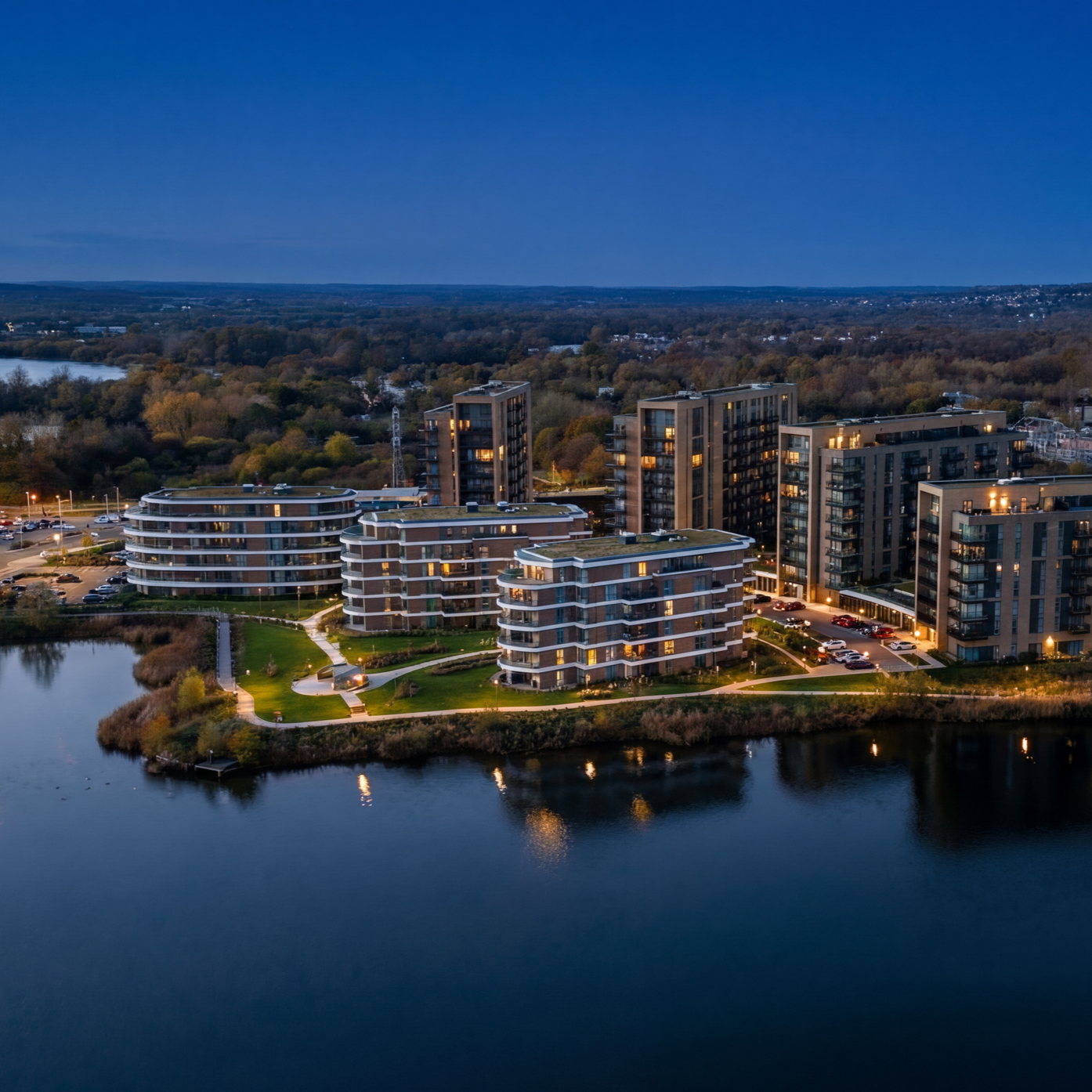 Nighttime aerial view of modern apartment buildings along a waterfront, with trees and a distant landscape in the background.