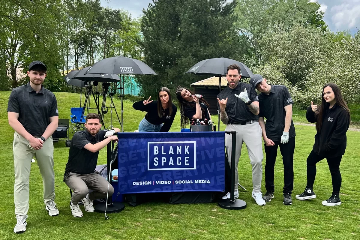 Group of seven people outdoors on a grassy field, with some holding umbrellas and others posing, standing behind a table with a "Blank Space" banner advertising design, video, and social media services.