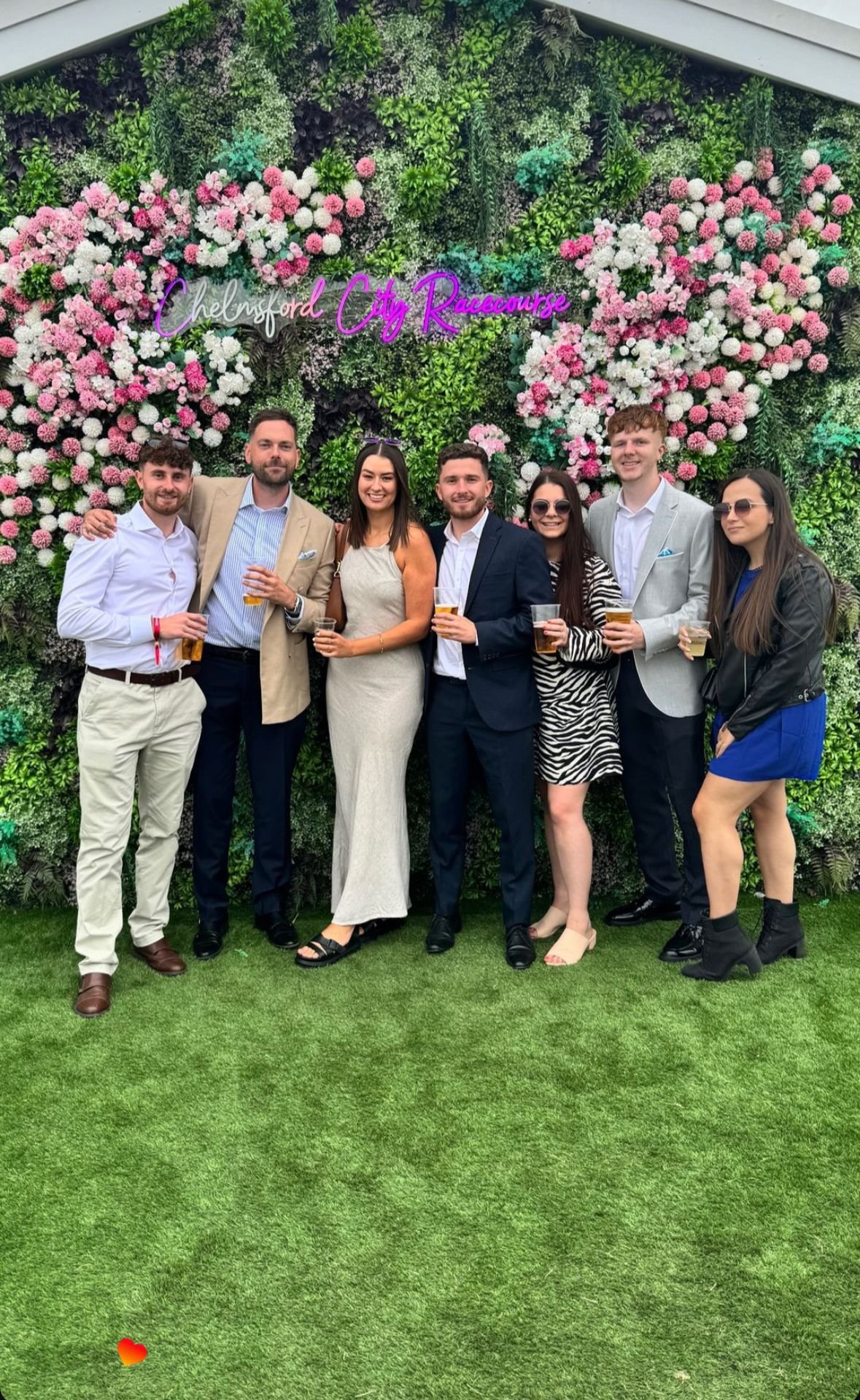 Group of seven people posing in front of a floral backdrop at Chelmsford City Racecourse, holding drinks, dressed in formal and semi-formal attire.