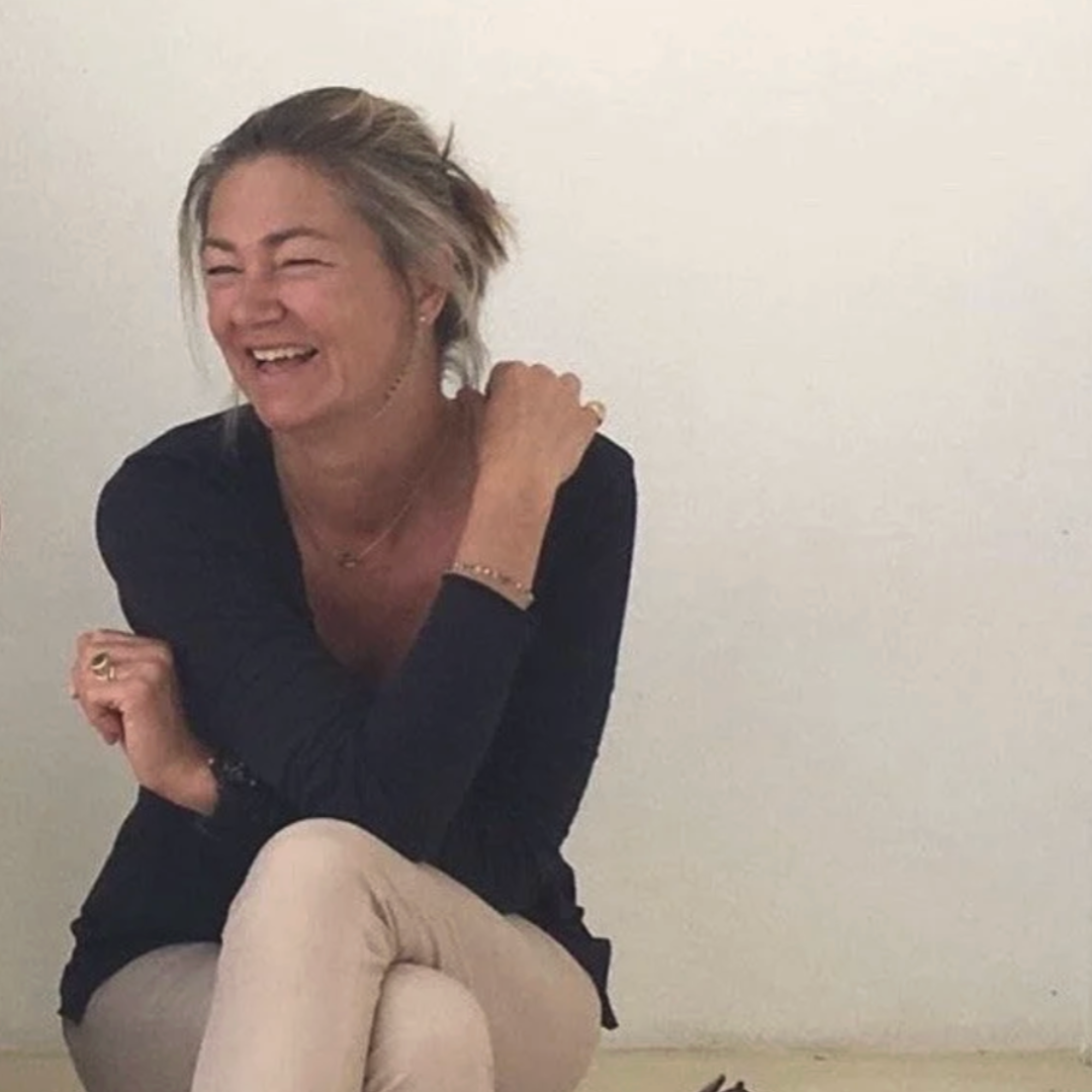 Une femme riante avec des cheveux blonds courts, portant un haut noir et un pantalon beige, assise contre un mur blanc.