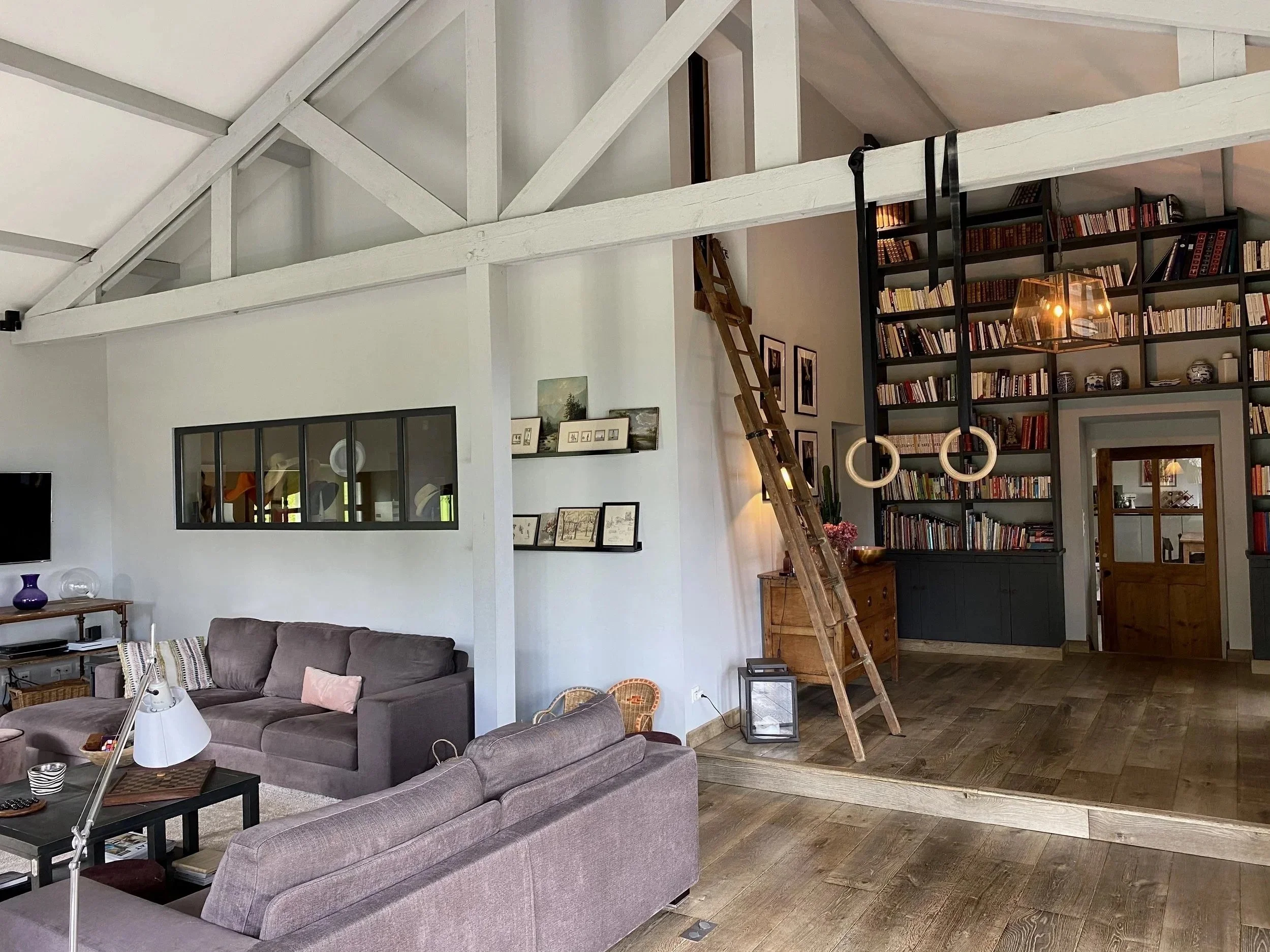 Salon contemporain avec canapés gris, une bibliothèque haute remplie de livres, un escalier en bois menant à une mezzanine, et des éléments décoratifs modernes.