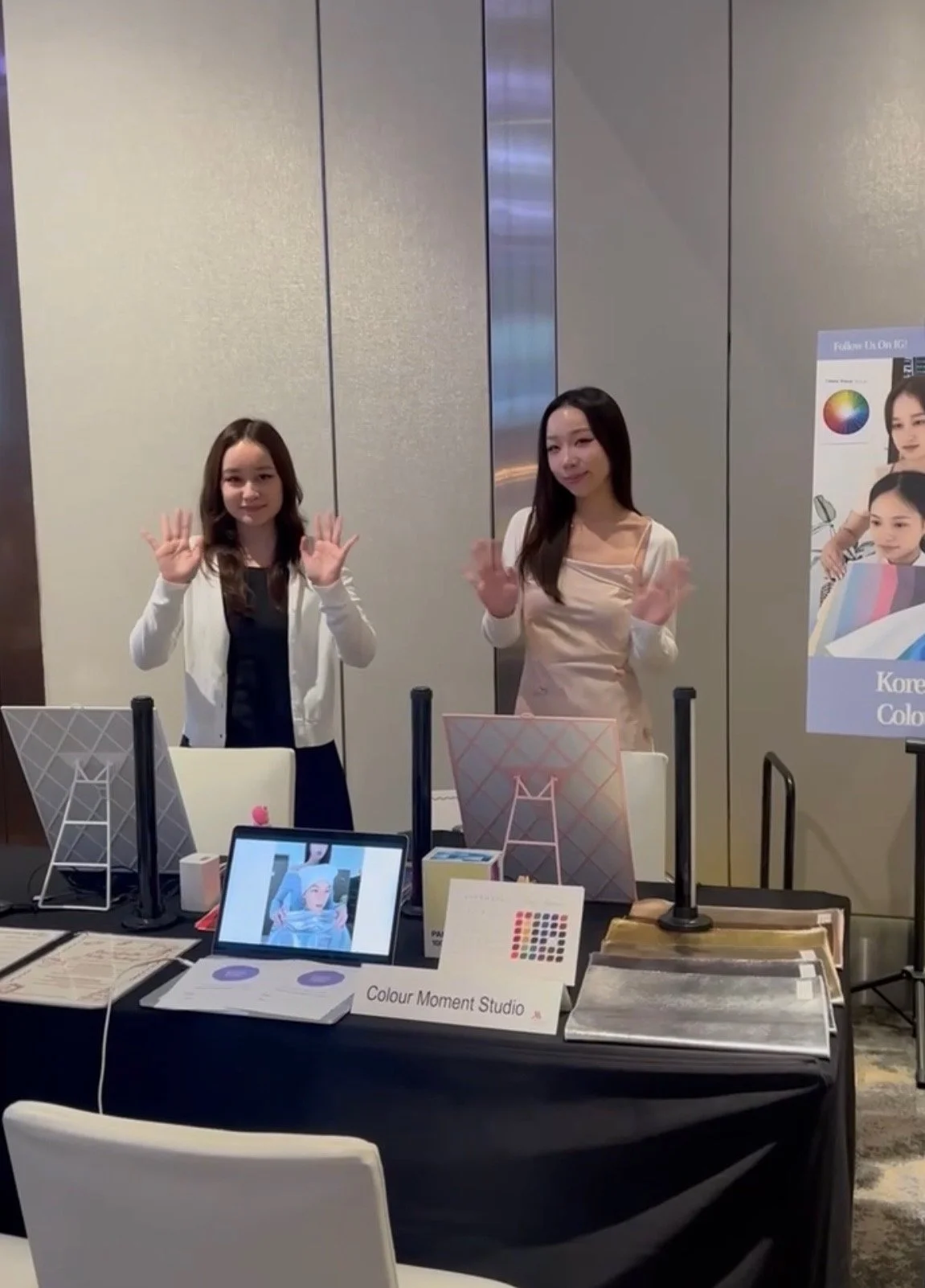 Two women standing behind a display table at a promotional event, waving at the camera. The table has various promotional materials, a tablet showing a woman with colorful makeup, and signs that read 'Colour Moment Studio' and 'Korean Colour'.